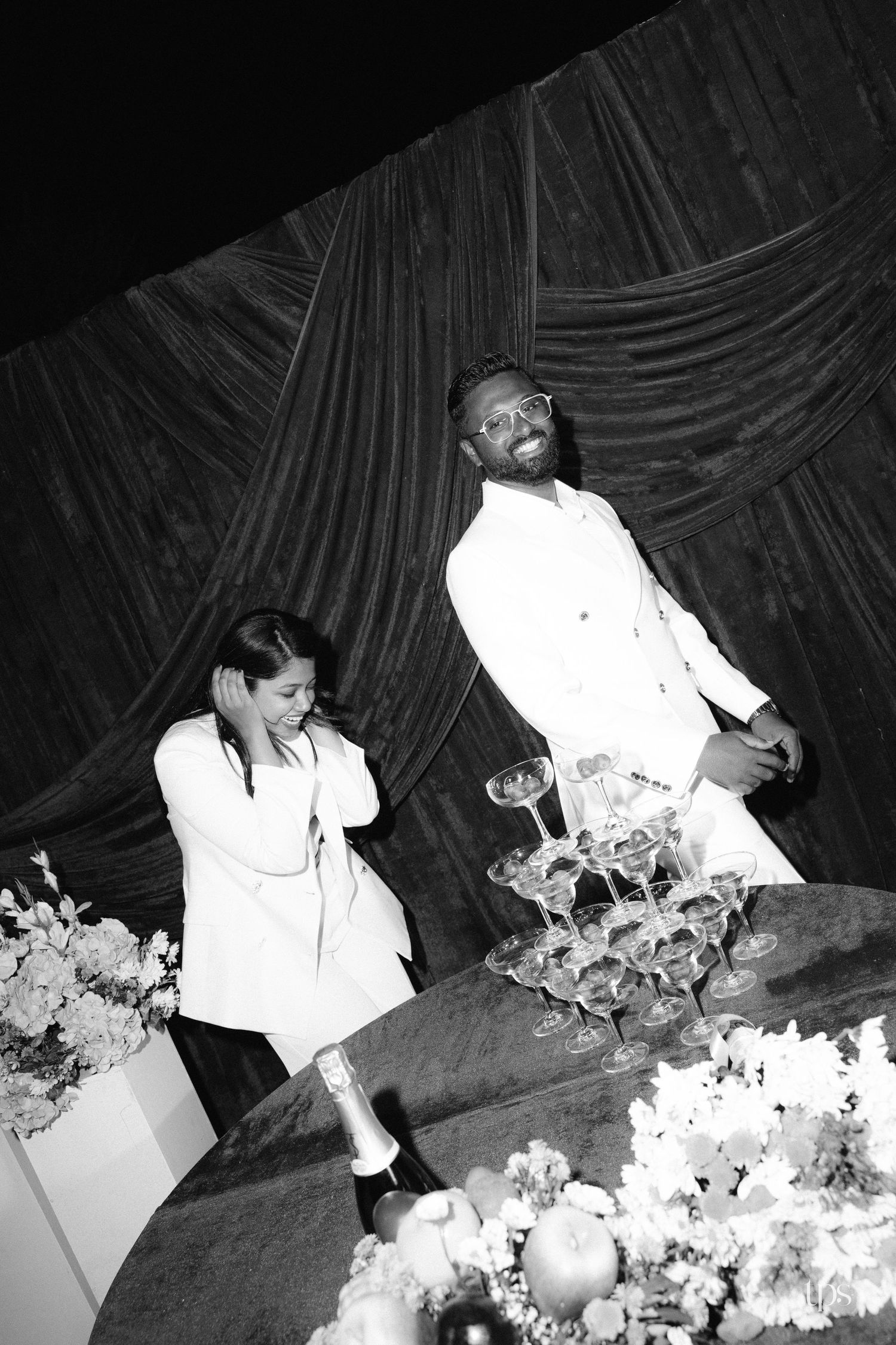 Black and white photo of two people in white suits laughing beside a champagne coupe tower, with draped curtains and flowers on a table.