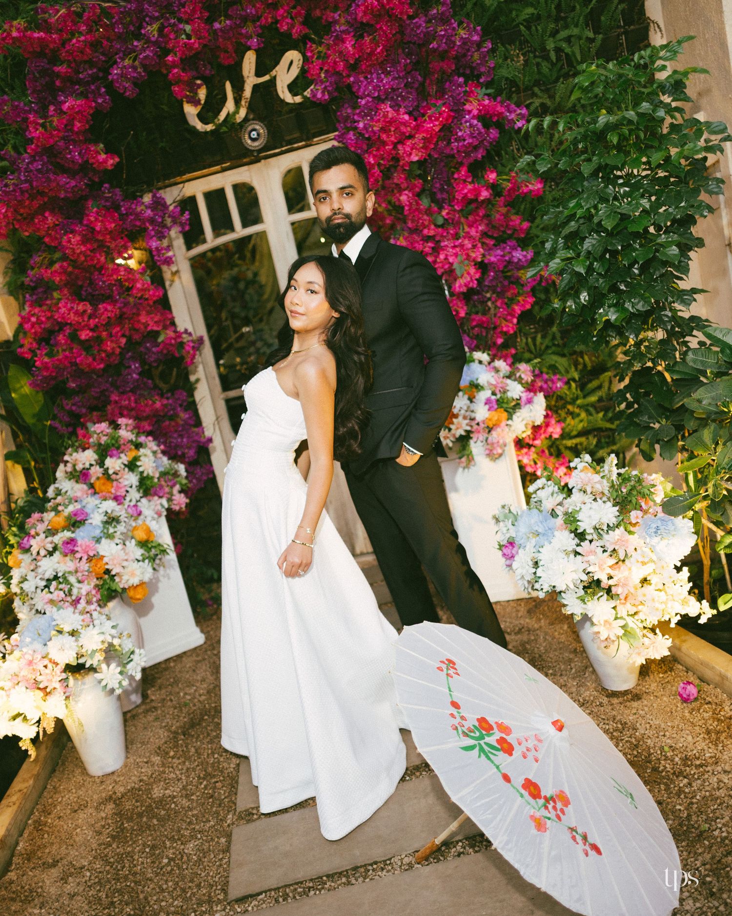 Bride in a white gown and groom in a black suit pose beside pink bougainvillea and floral arrangements with a painted paper parasol.