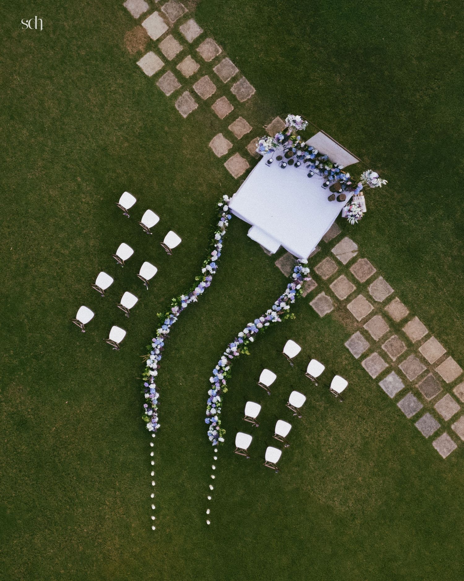 Aerial view of an outdoor wedding ceremony setup with a white platform, floral arch, blue flower garlands, and white chairs on green grass.
