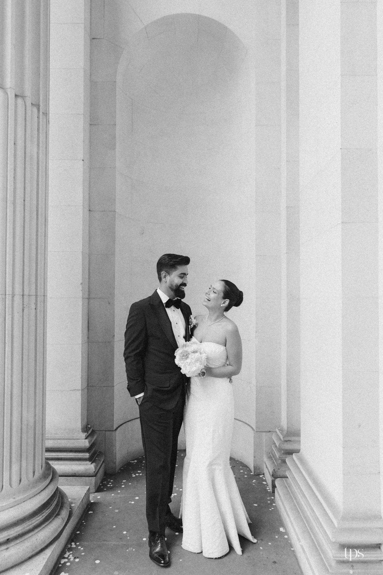 Black and white photo of a bride and groom sharing a joyful moment between grand columns at their elegant wedding venue.