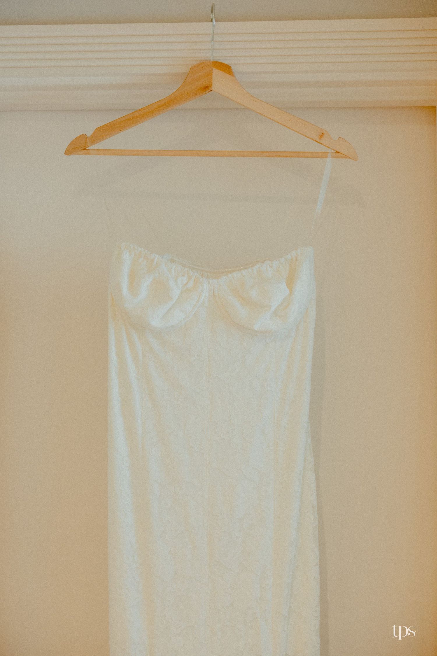 A strapless white lace wedding dress hanging on a wooden hanger against a cream wall.