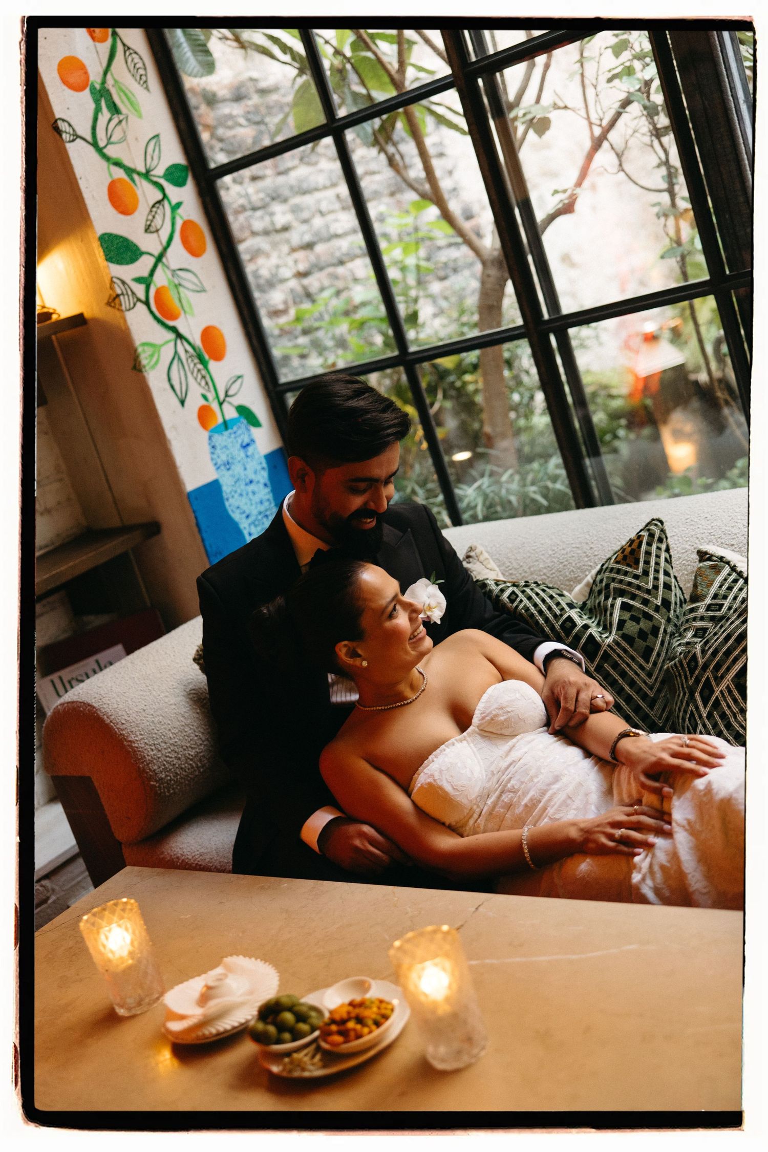 Couple in wedding attire relaxing on a couch by large windows with candles and snacks on table.