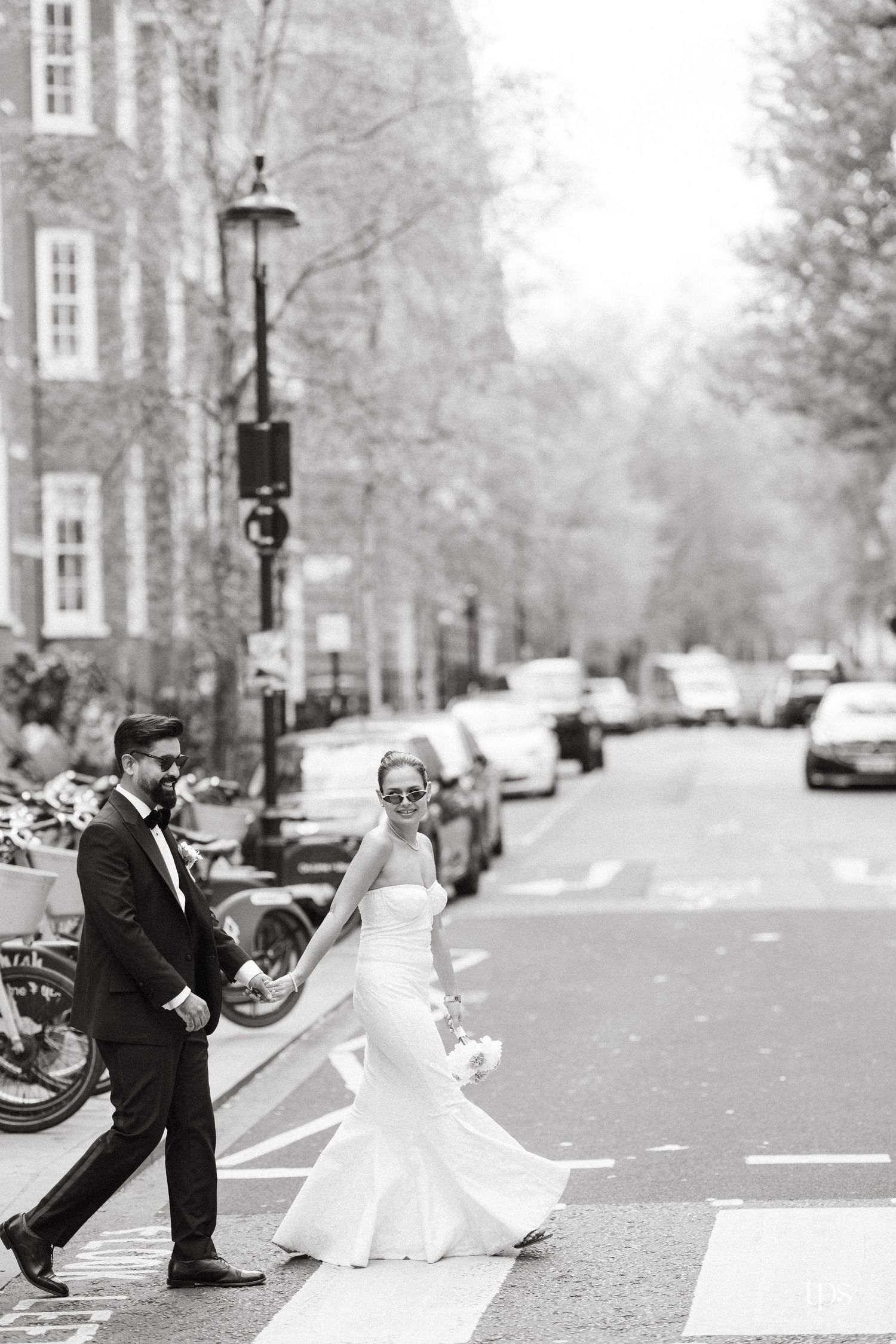 Black and white photo of bride and groom holding hands crossing a street in elegant wedding attire.