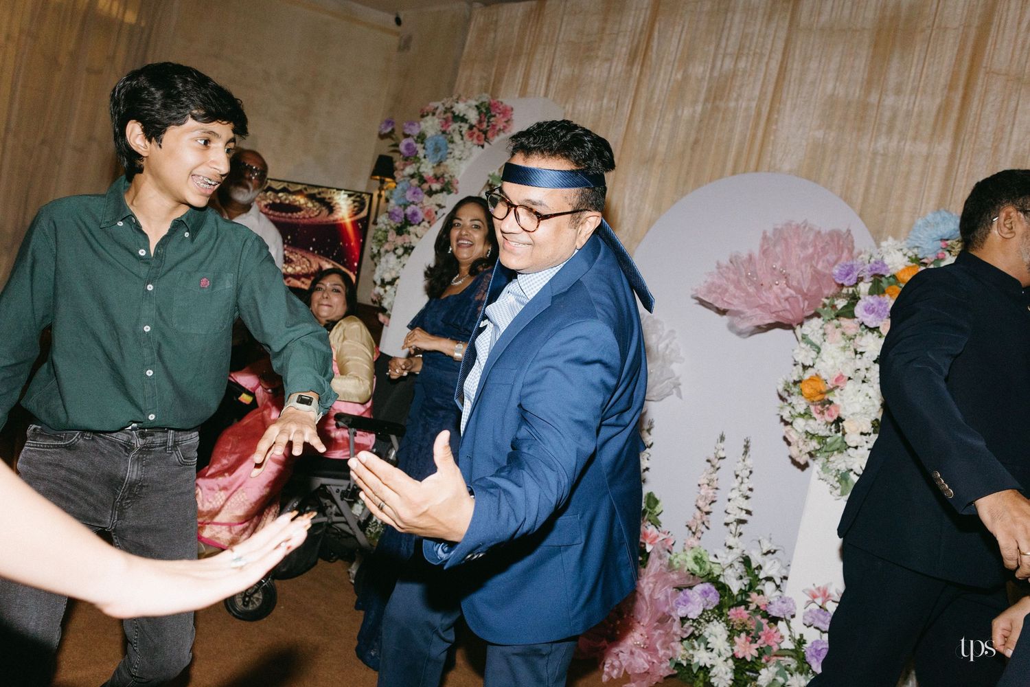 Guests dance and celebrate at a festive wedding reception, with a man in a blue blazer and headband smiling beside floral decorations.