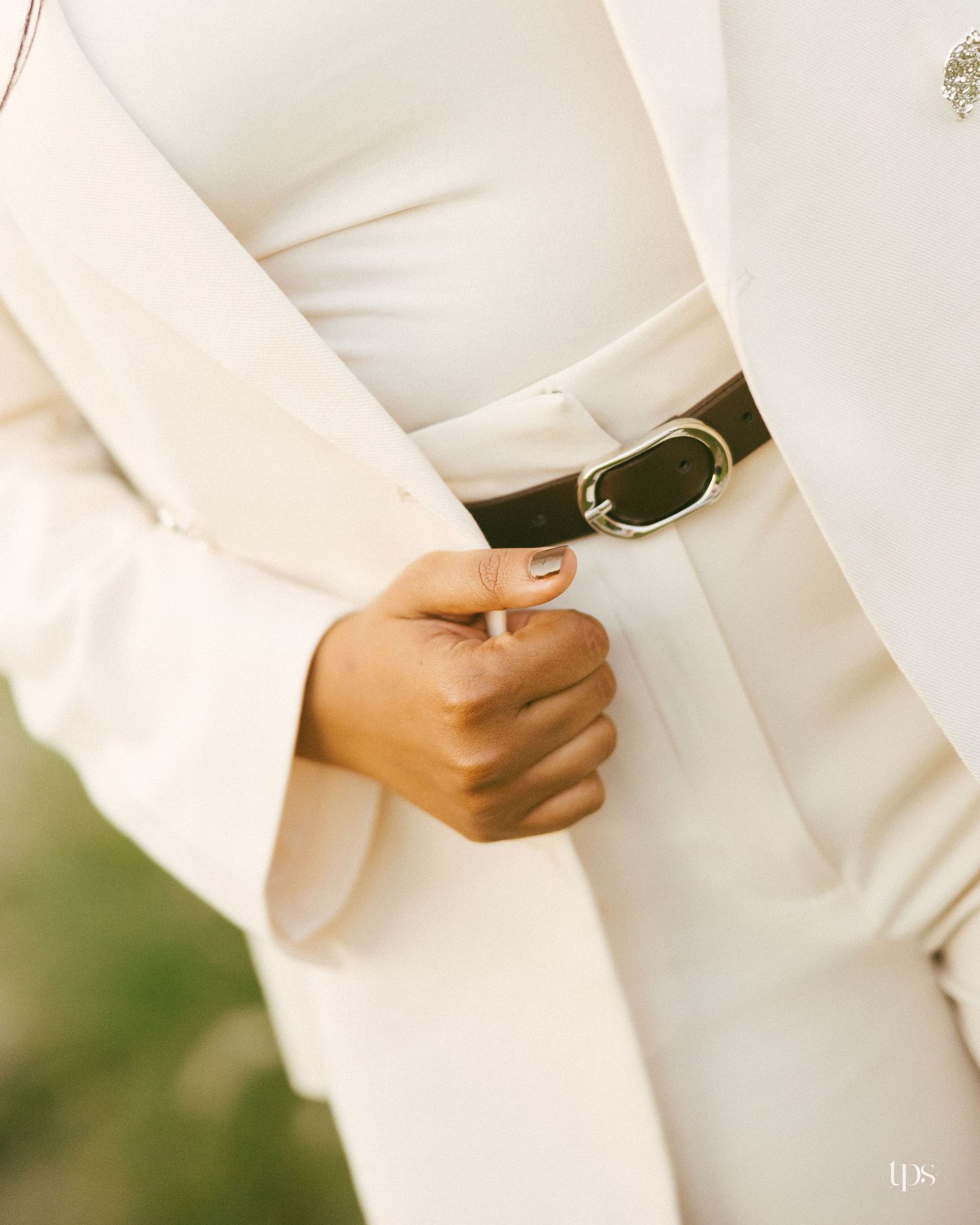 Woman in a cream blazer and matching trousers with a black leather belt and gold buckle, hand resting at her waist outdoors.
