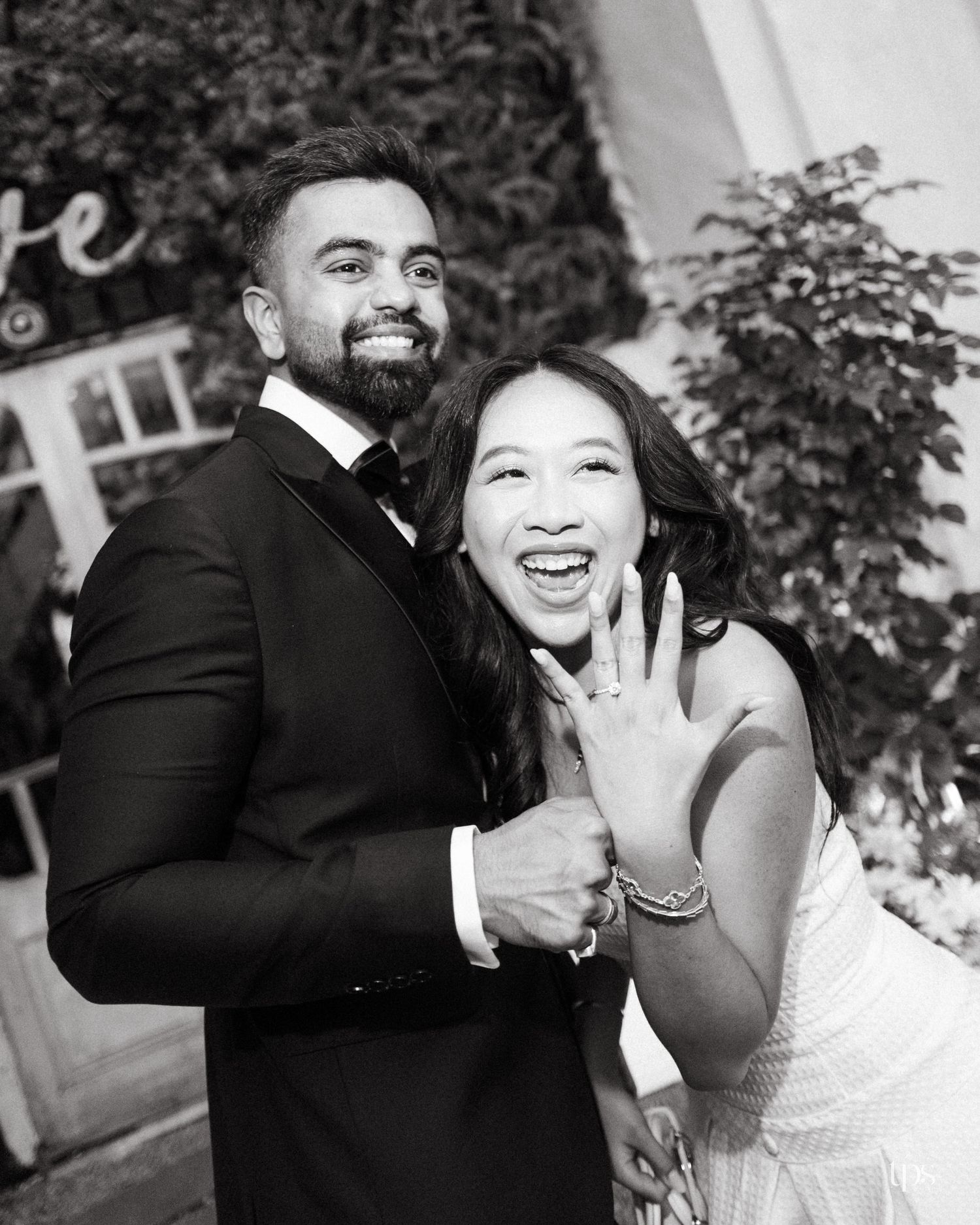Smiling couple in a black-and-white engagement photo as the excited woman shows her ring beside a bearded man in a tuxedo.