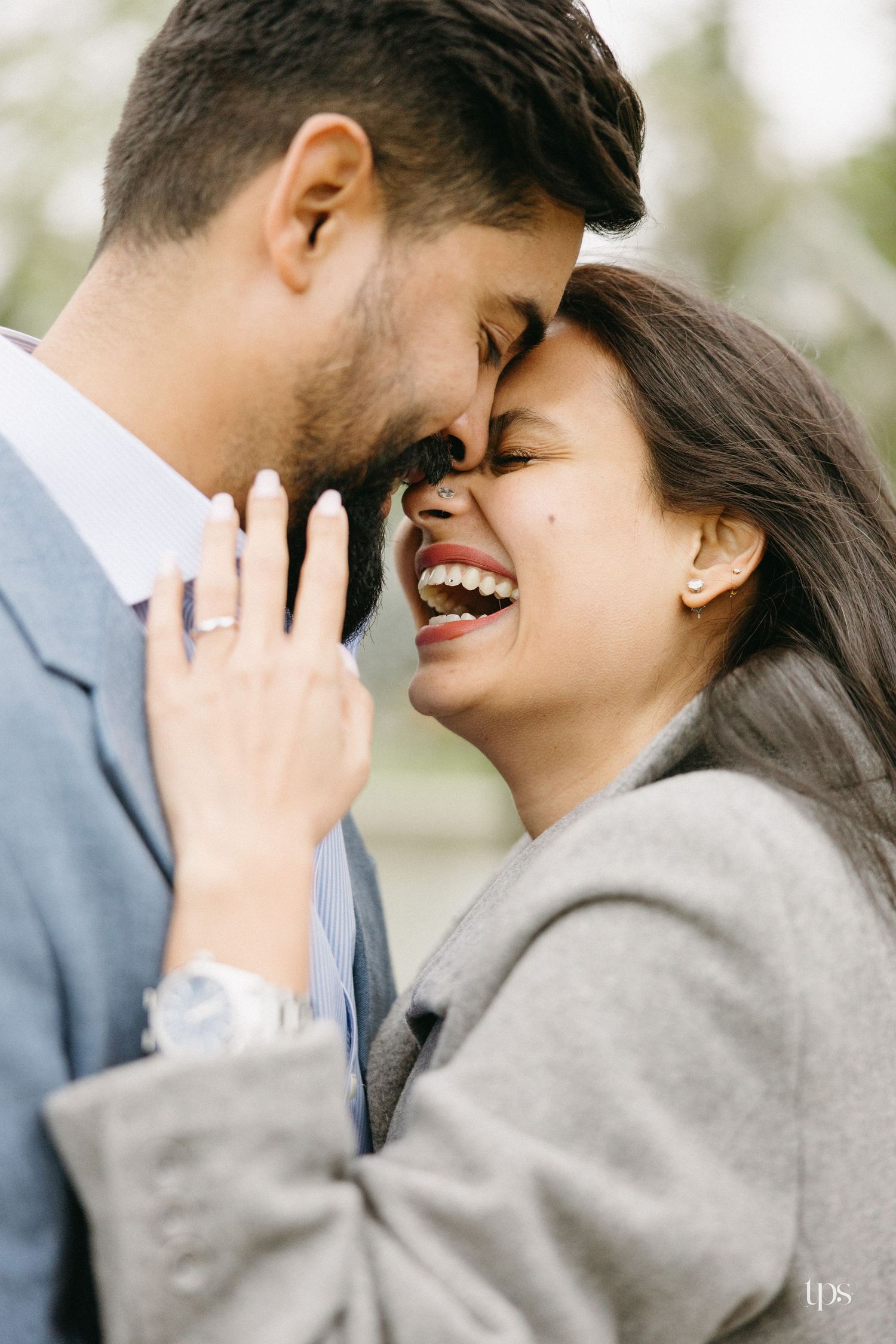 A joyful couple in gray suits share an intimate forehead-to-forehead moment, the woman laughing with an engagement ring visible.