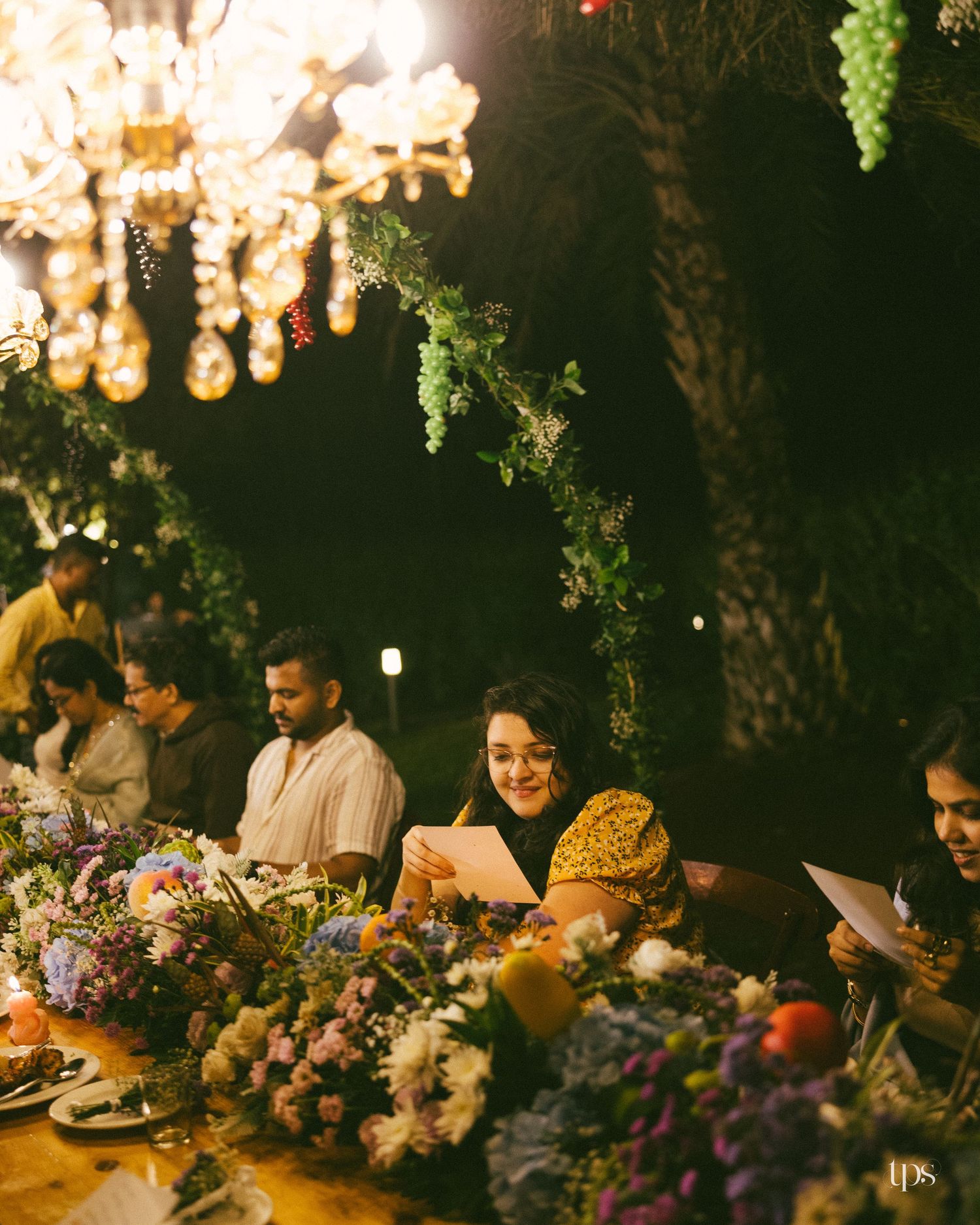 Guests holding papers sit at a flower-covered outdoor dinner table under a glowing chandelier and greenery at night.