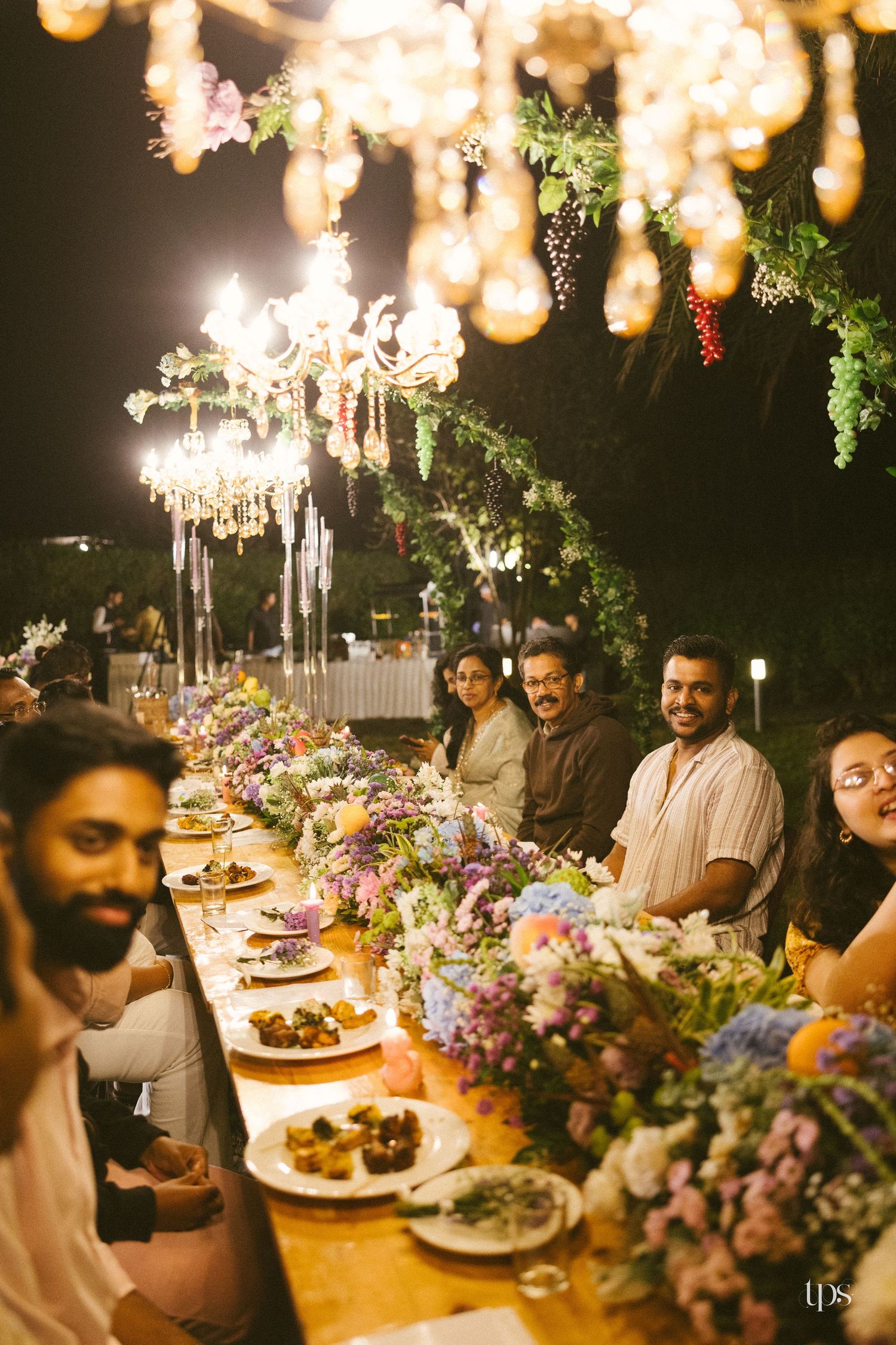 Guests smile at a long outdoor wedding dinner table decorated with colorful flowers, plated food and glowing chandeliers at night.