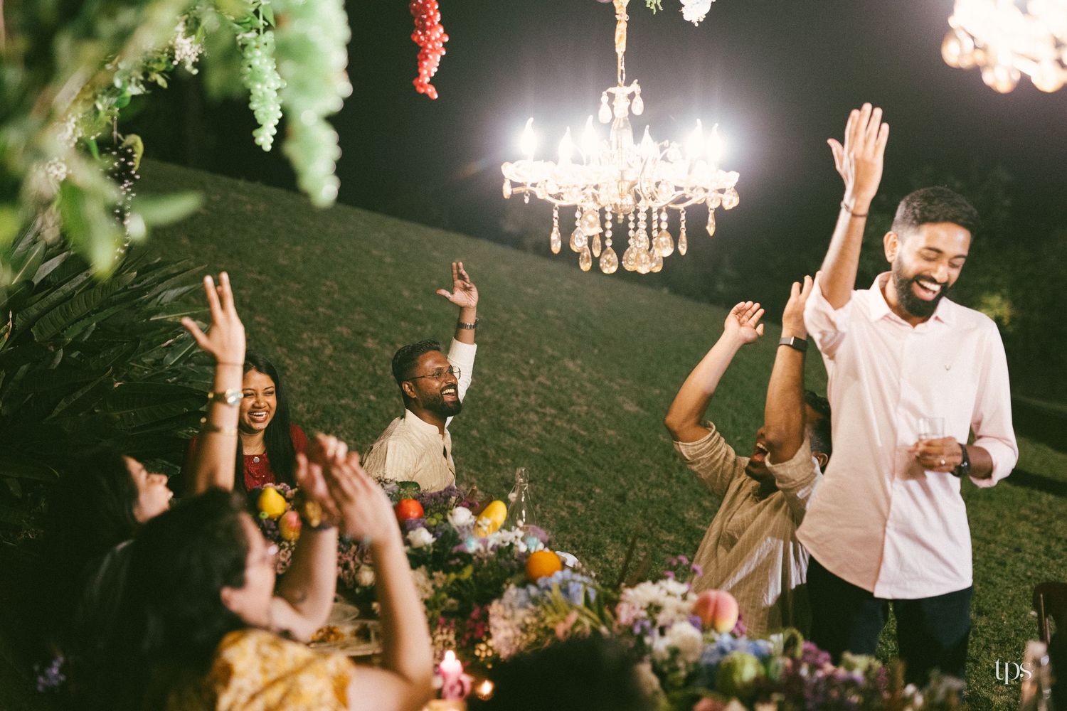Smiling guests raise their hands and cheer around a flower-decorated table at an outdoor evening party under a glowing chandelier.