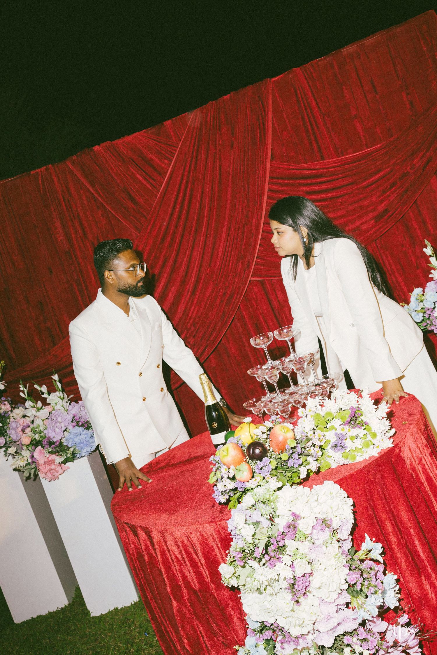Two people in white suits stand facing each other across a red velvet table with flowers, fruit, and a champagne tower.