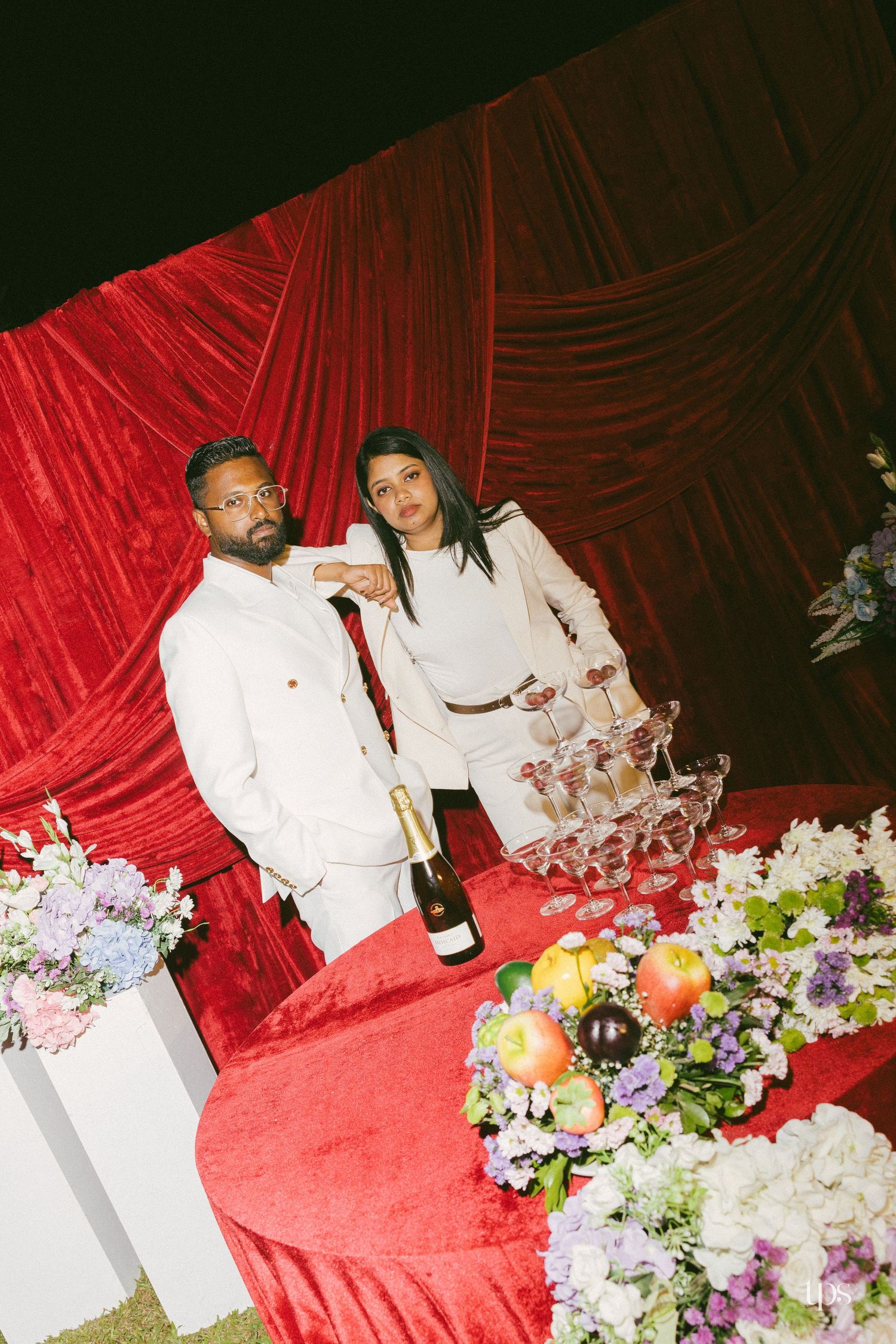 Couple in white outfits poses beside a champagne tower and bottle on a red velvet table with fruit and flowers against a red drape.