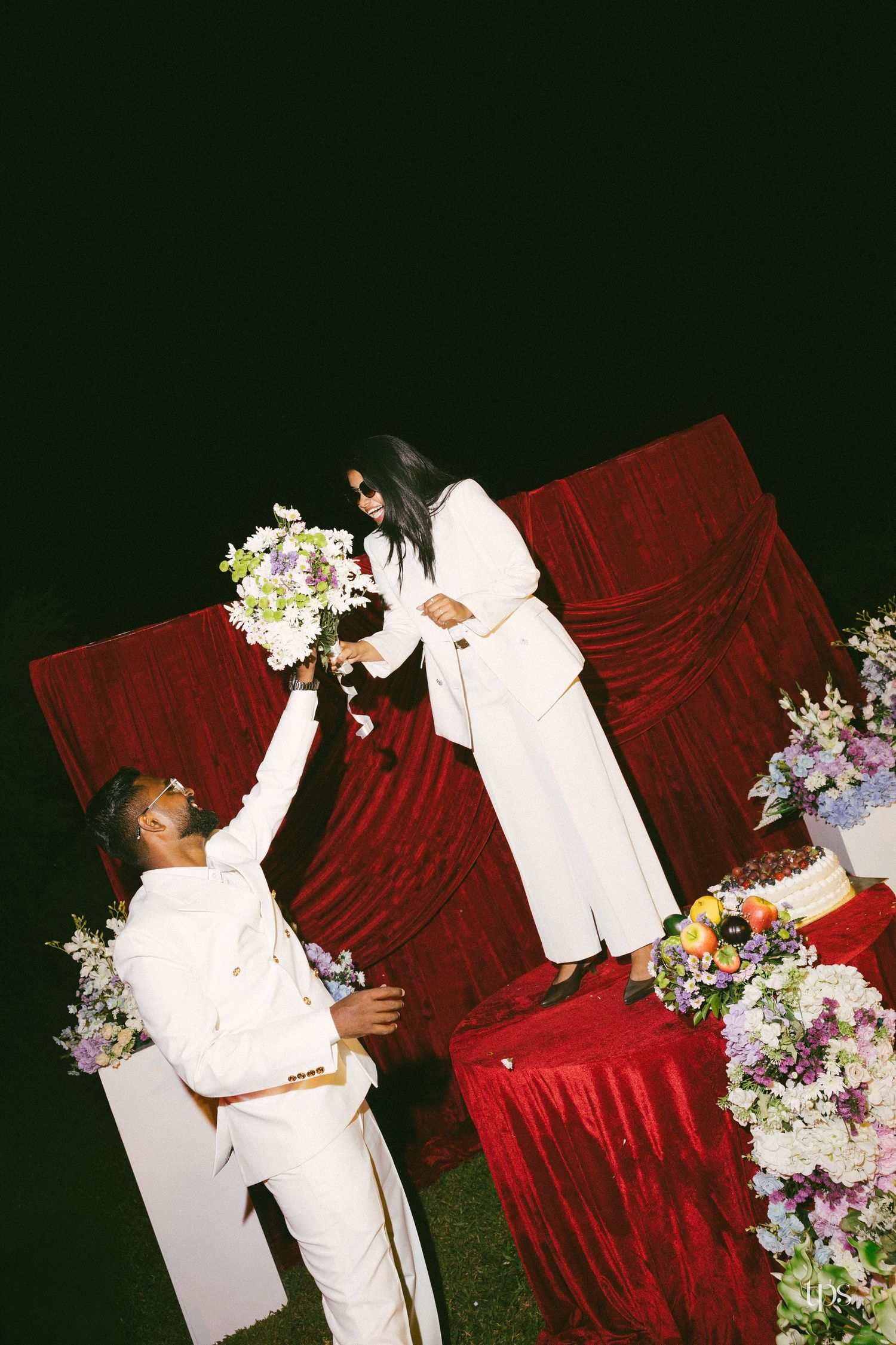 Couple in white suits at a night wedding celebration, holding a floral bouquet near a red velvet backdrop and a cake table.