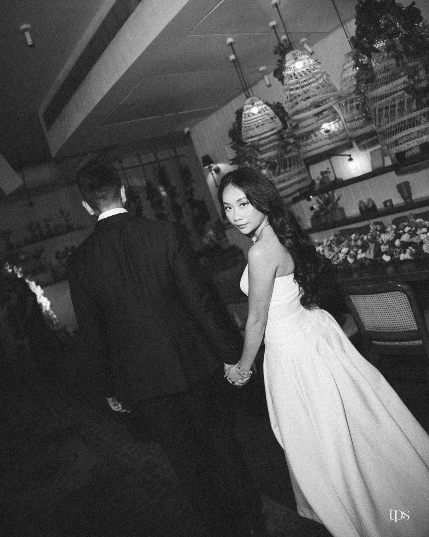 Black and white photo of a bride in a strapless white gown holding hands with a groom in a dark suit inside an elegant restaurant.