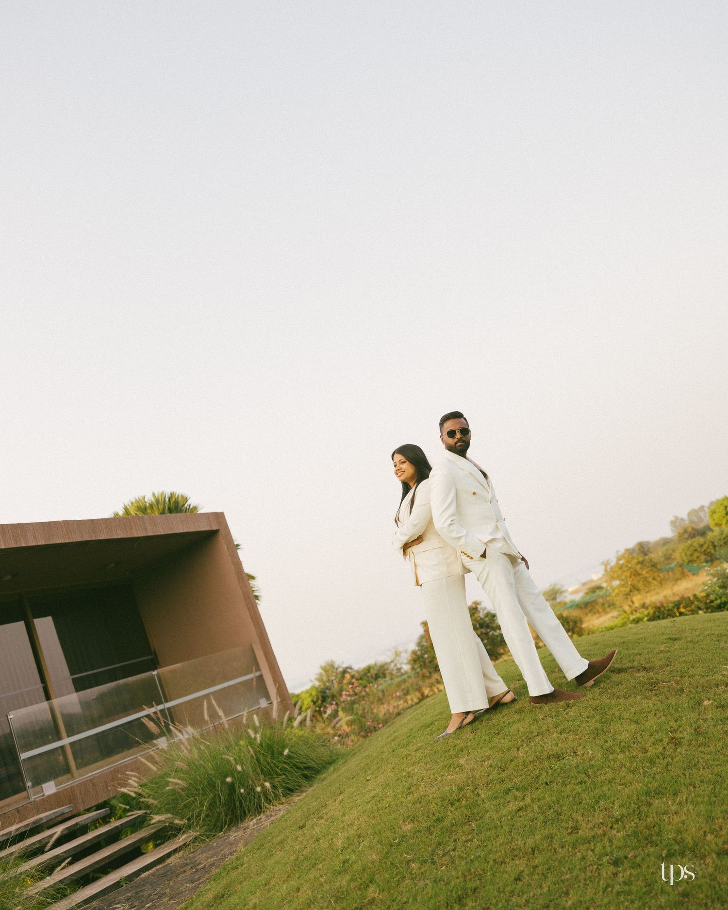 Couple in matching white suits stand back to back on a grassy hillside beside a modern building under a soft hazy sky.