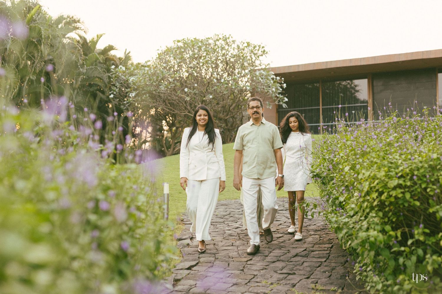 Family of three in white outfits walking along a stone paved garden path lined with purple flowers and lush greenery near a modern building.