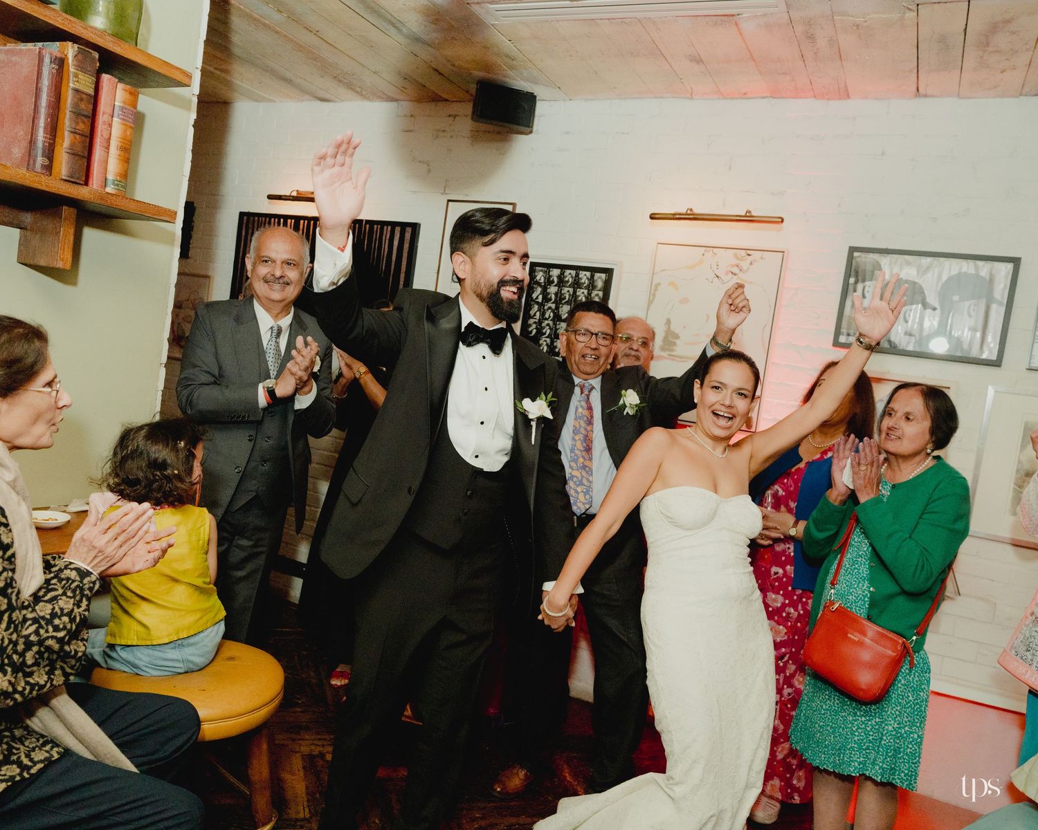 Bride and groom hold hands celebrating joyfully with smiling guests clapping at an intimate indoor wedding reception.