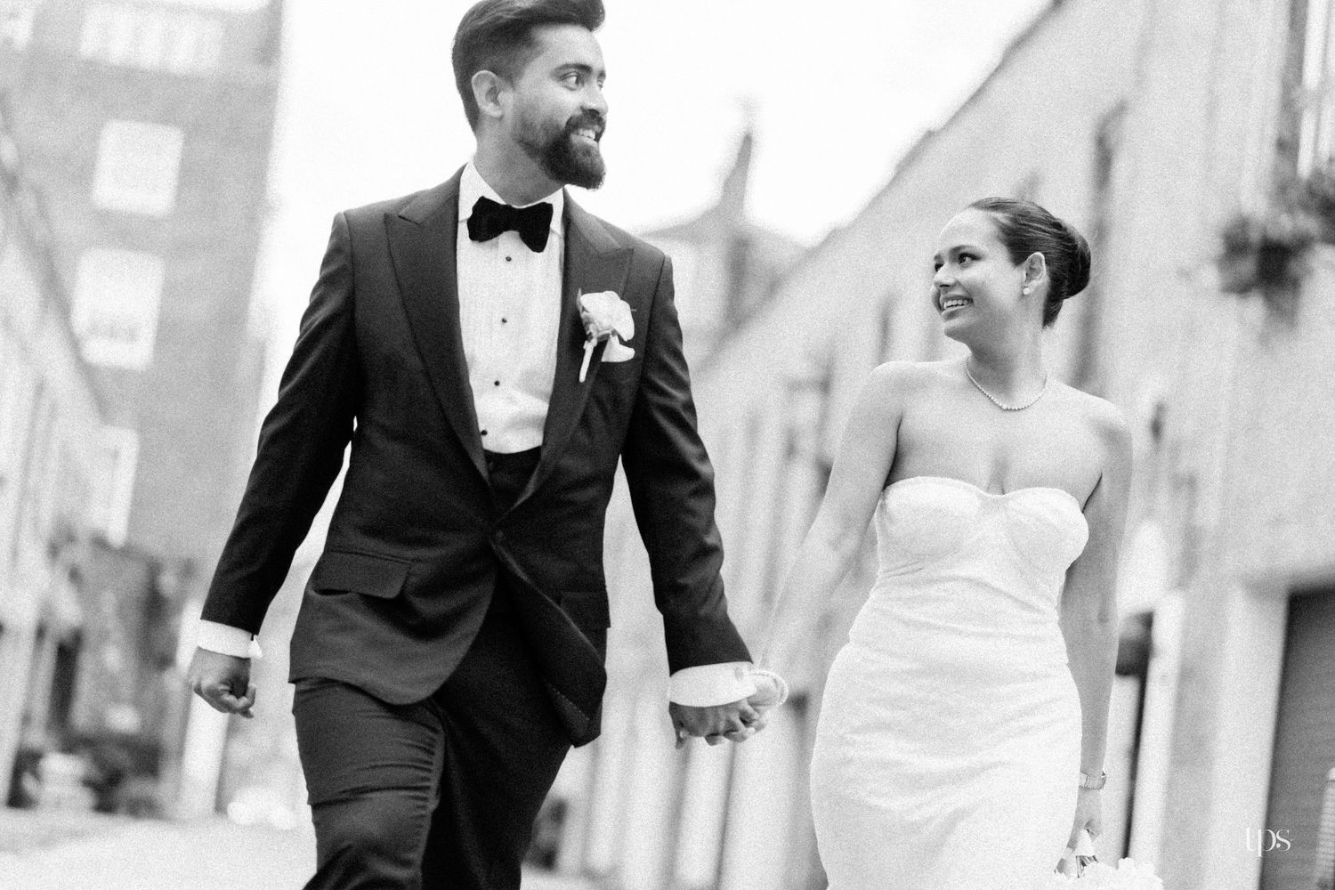 Black and white photo of a smiling bride and groom holding hands walking together on a city street on their wedding day.