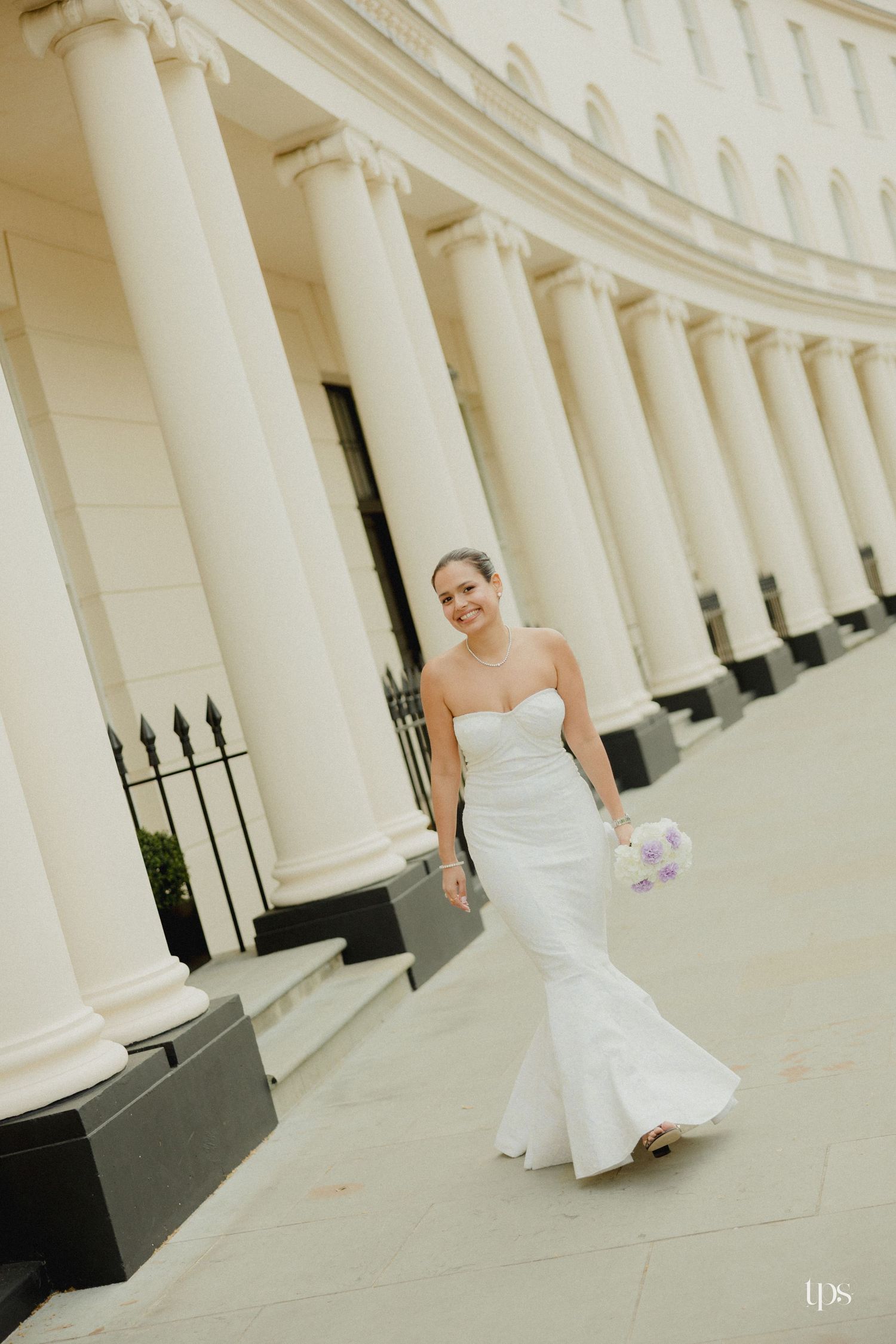 Bride in strapless mermaid gown walks past grand white columns holding purple bouquet at elegant venue.