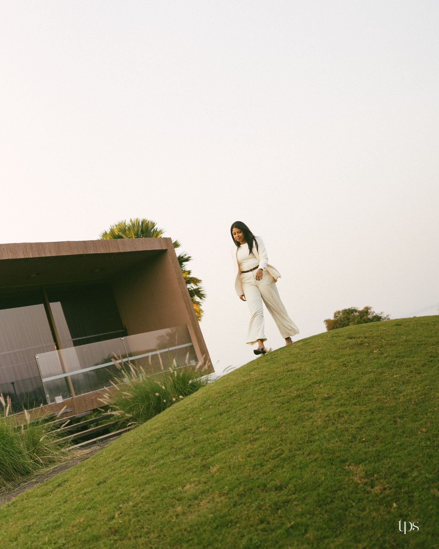 Woman in a white outfit walks along a grassy hill beside a modern brown building with glass railings under a soft pastel sky.