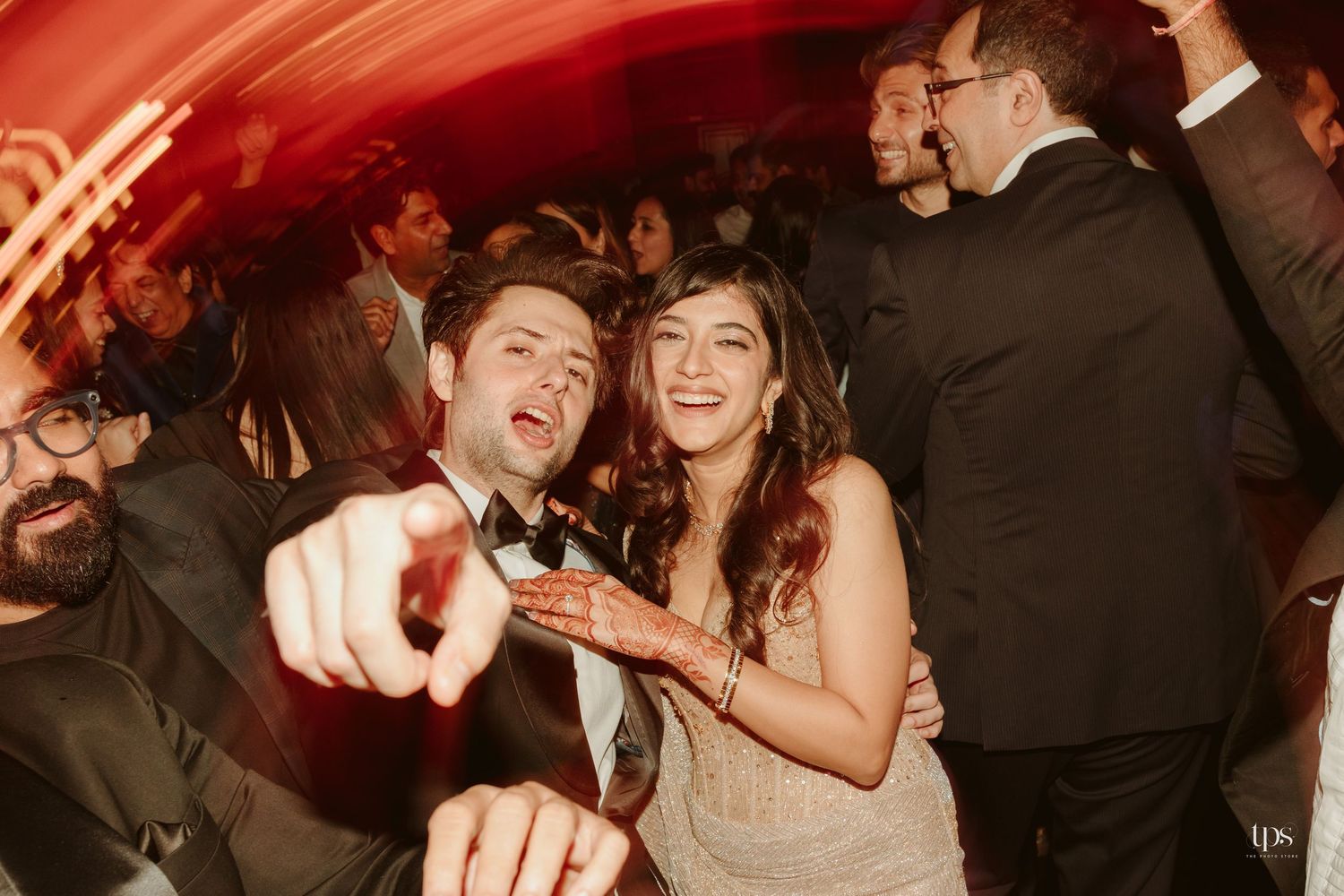 Couple dancing joyfully at wedding reception, groom in tuxedo pointing at camera, bride in white dress smiling, crowded dance floor.