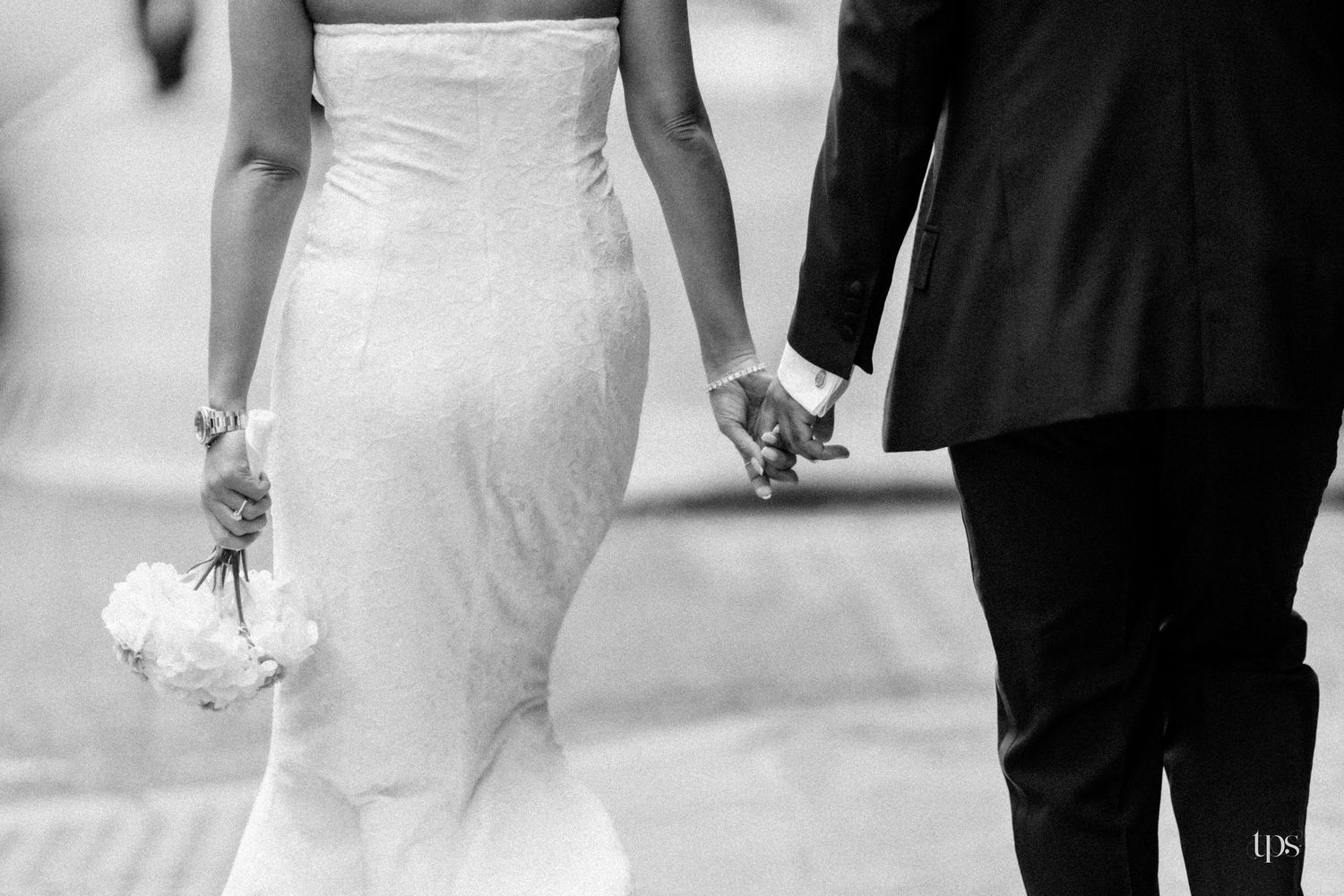 Black and white photo of a bride in a fitted gown holding flowers and groom in a suit walking hand in hand.