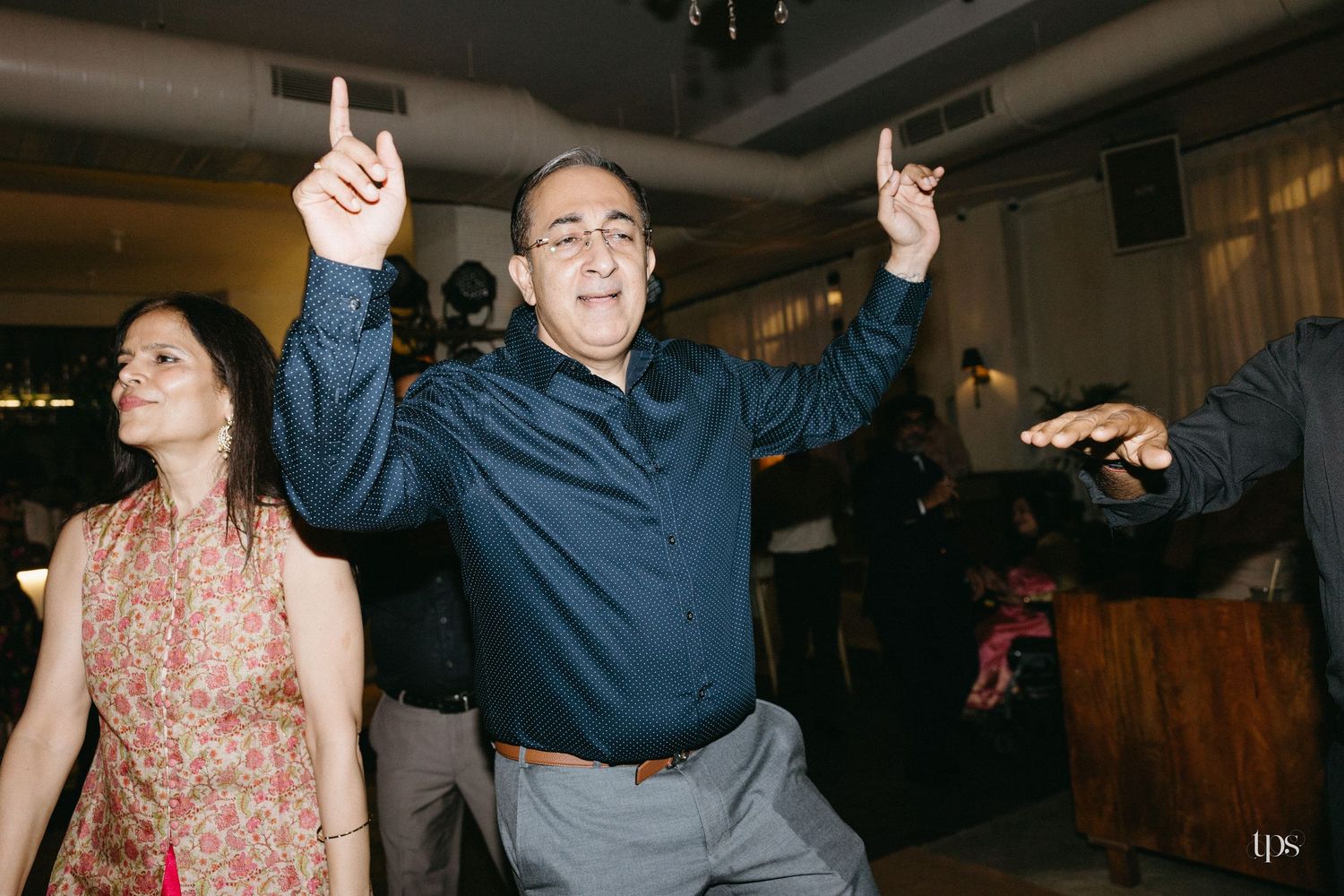 Smiling man in a blue shirt and glasses dances with raised hands at a lively indoor party while a woman in a floral top looks on.