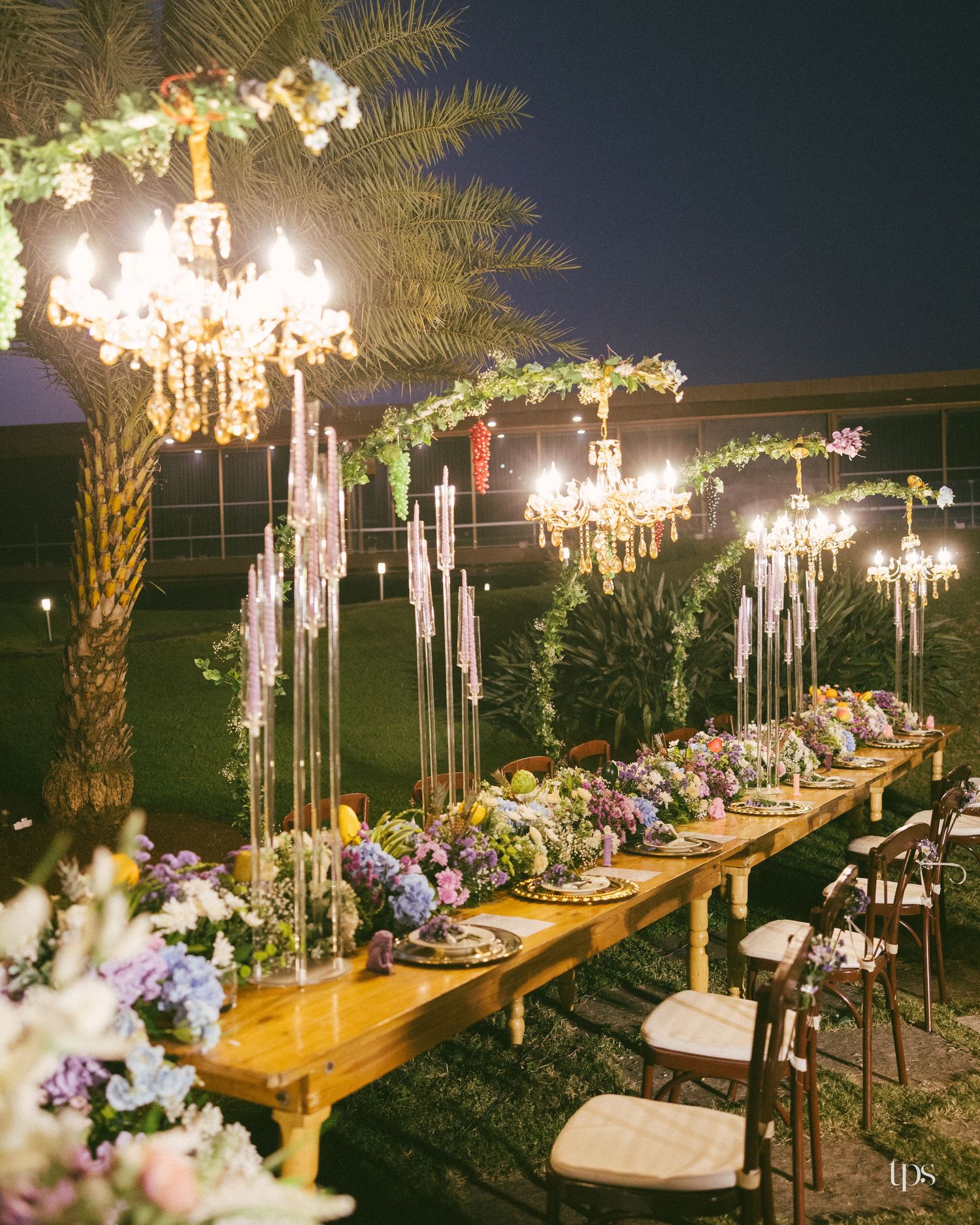 Elegant outdoor wedding reception table at night with crystal chandeliers, tall candles, pastel floral centerpieces and greenery garlands.