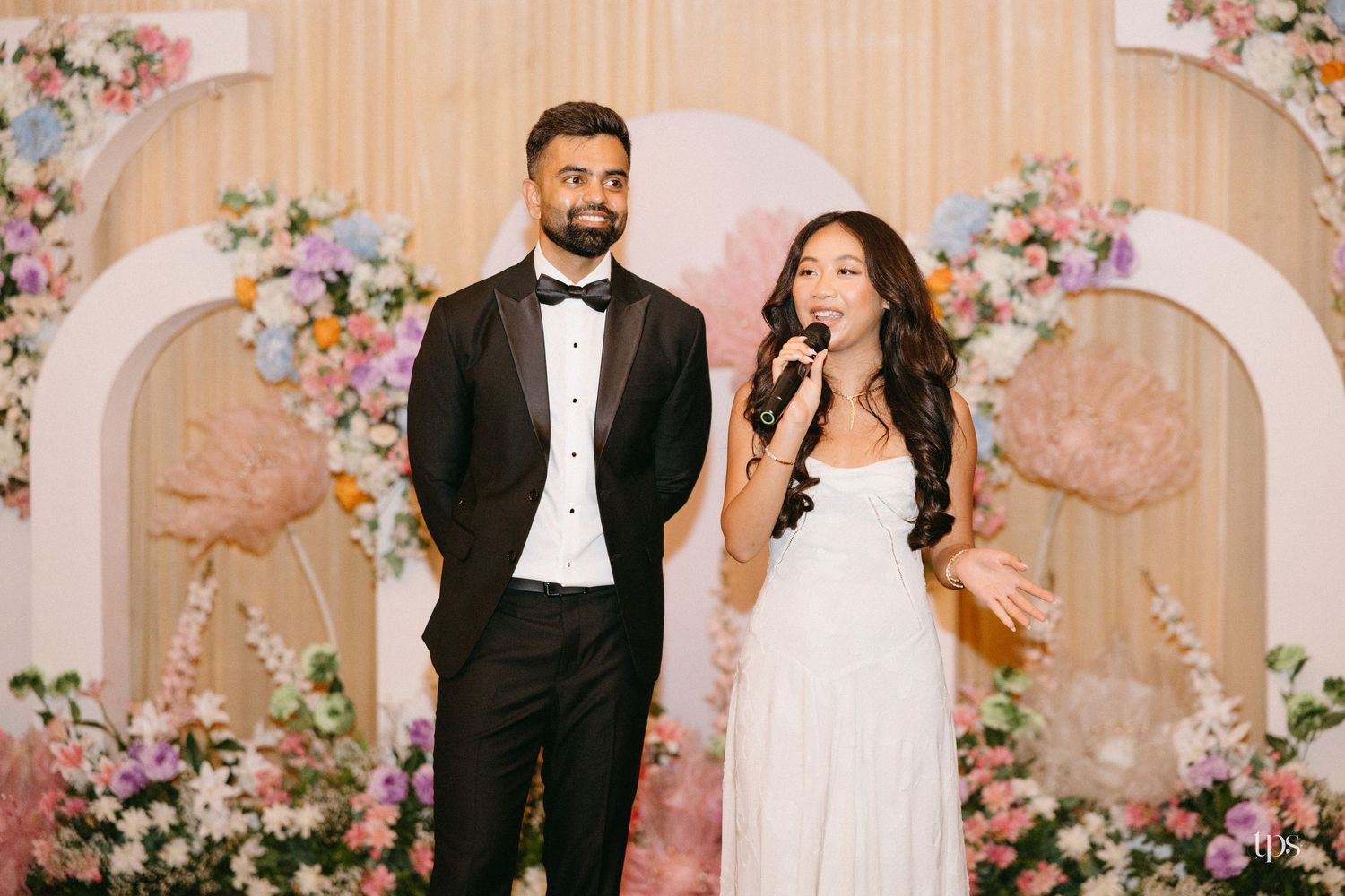 Man in a black tuxedo and bow tie stands beside a woman in a white dress speaking into a microphone at a wedding with colorful floral decor.