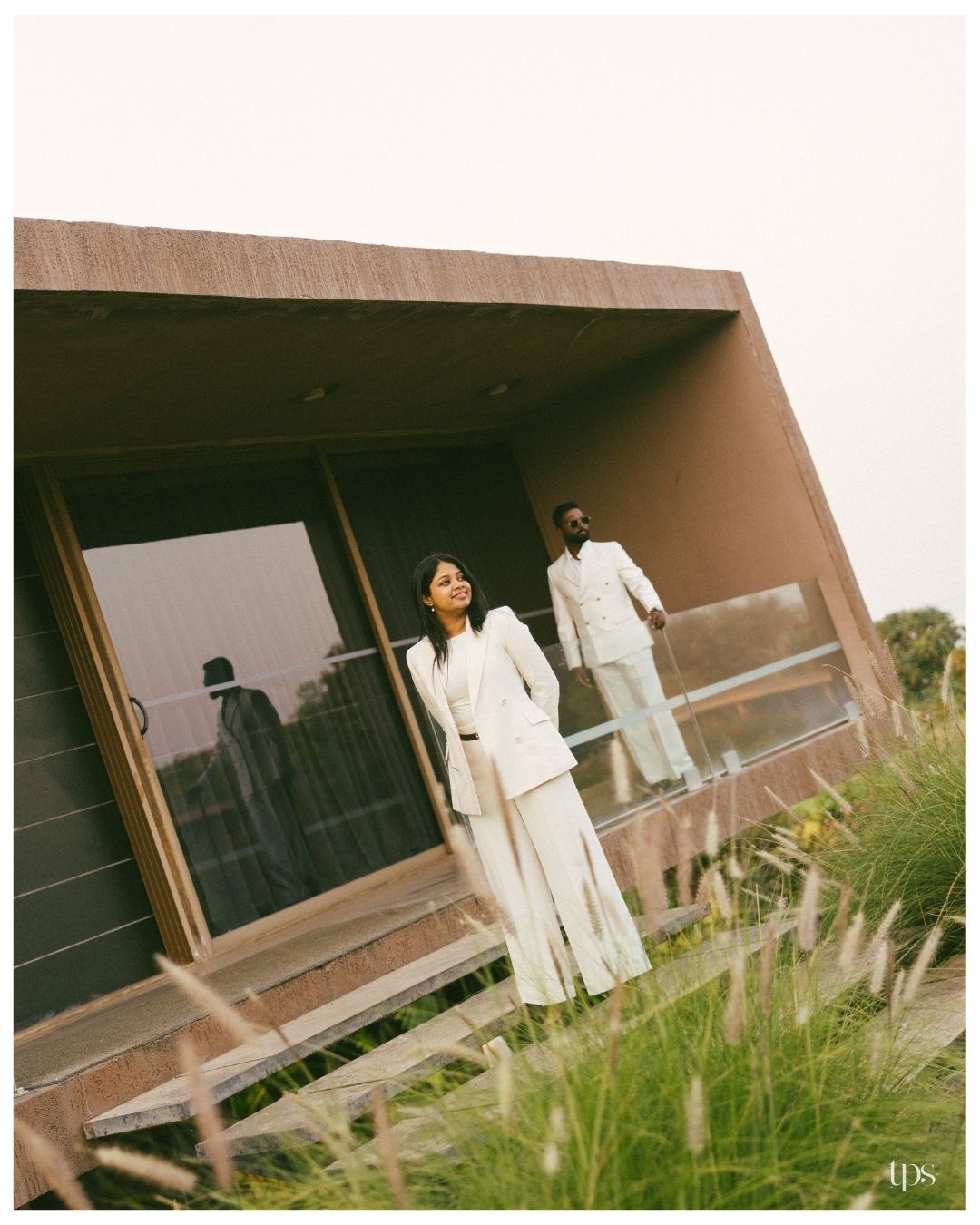 Couple in matching white suits pose on the balcony of a modern earth-toned building, framed by tall wild grass.