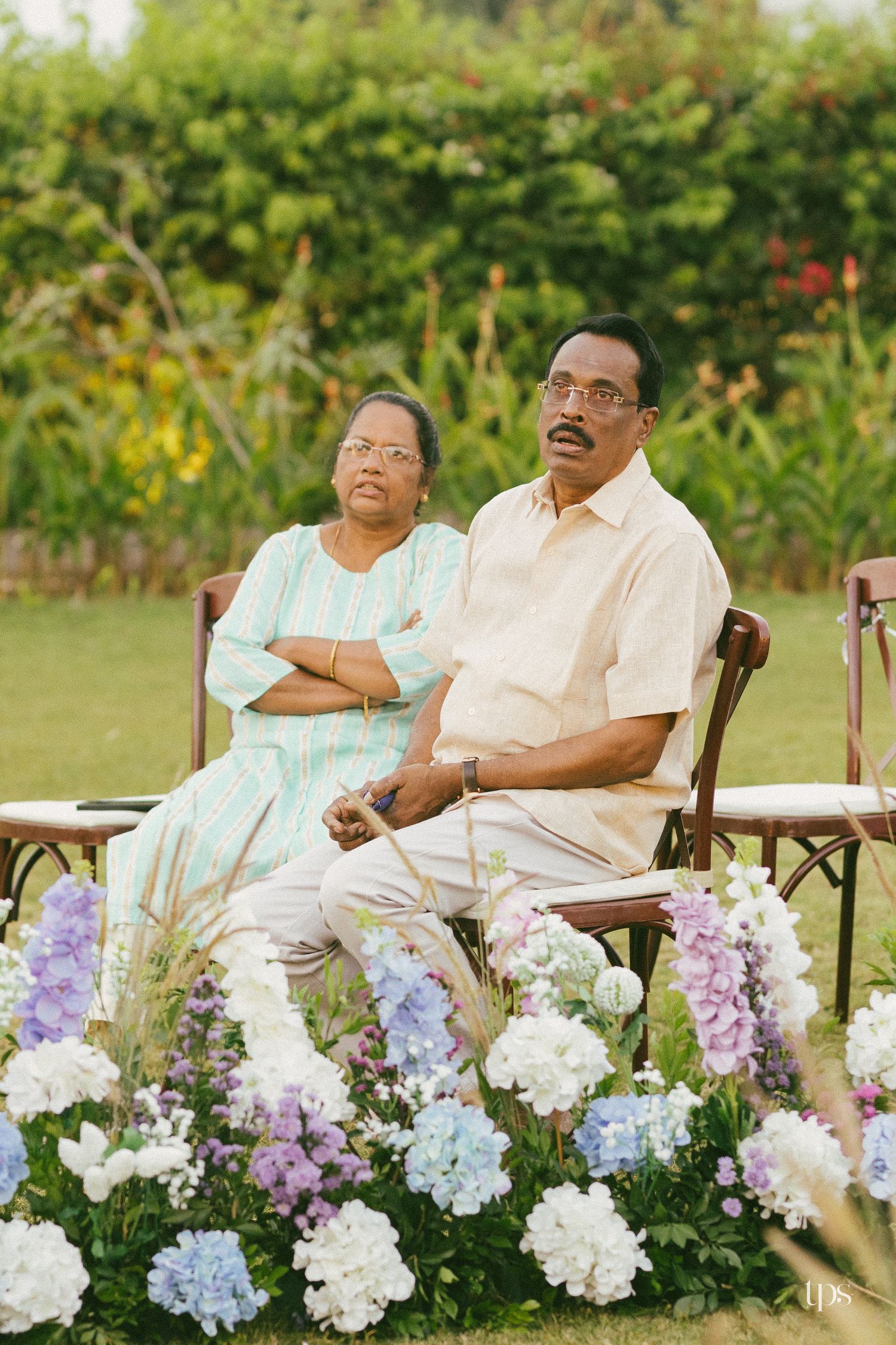 Elderly couple sitting side by side on wooden chairs in a sunlit garden, with blue and white hydrangeas blooming in the foreground.