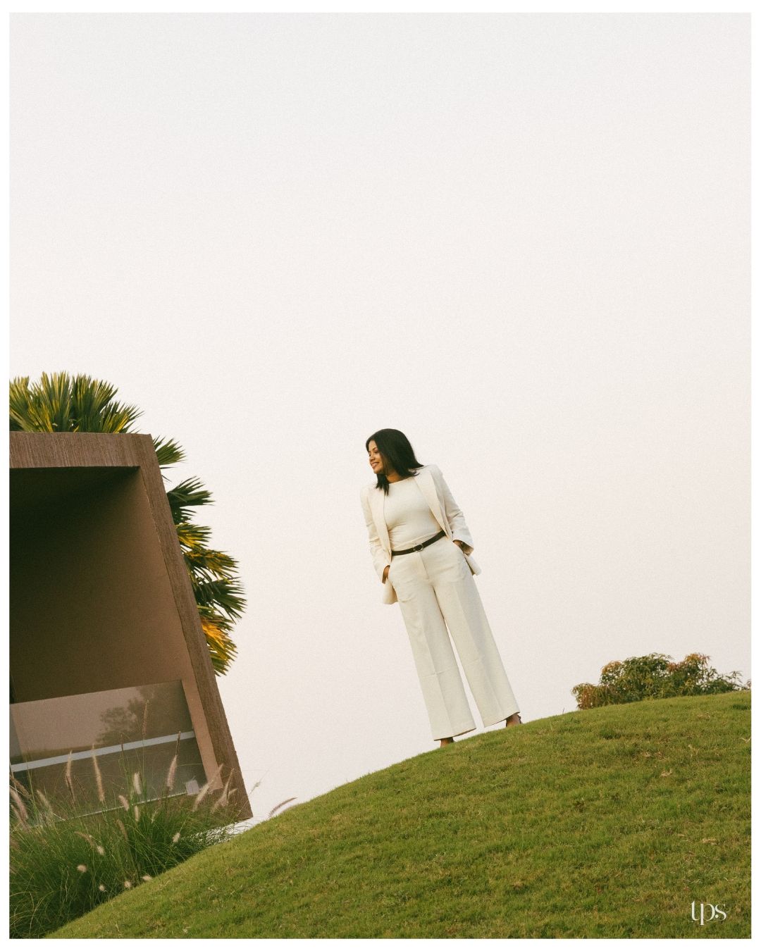 Woman in an all-white pantsuit with a brown belt stands on a grassy hill beside a modern angular building and palm tree under a hazy sky.