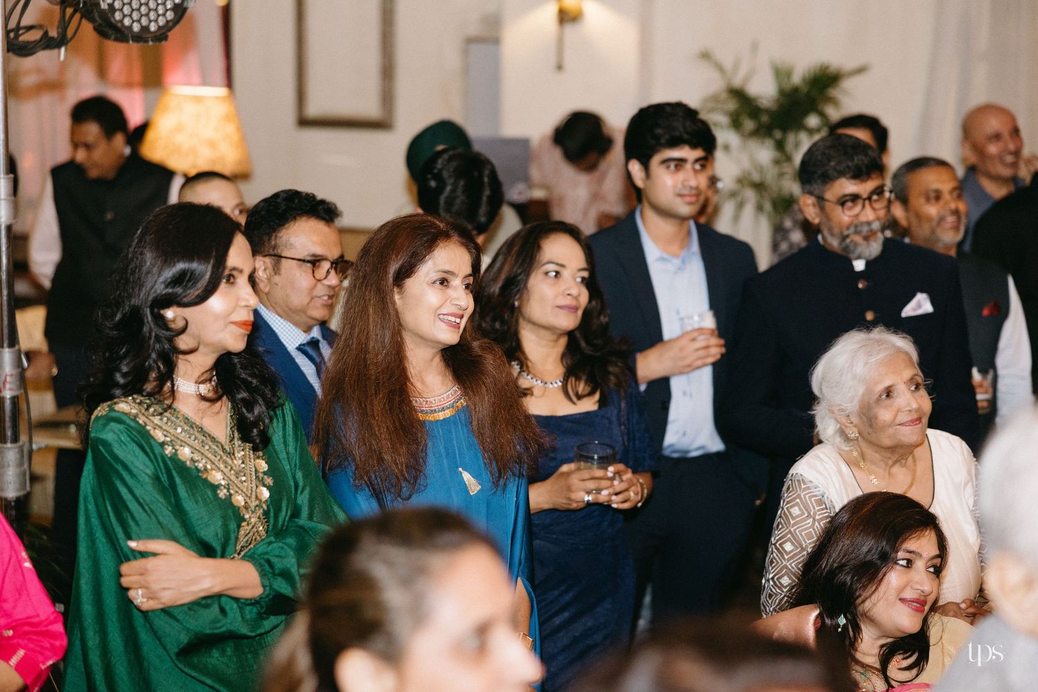 Guests in elegant formal attire stand and smile together at an indoor evening gathering, with warm lighting and a festive atmosphere.