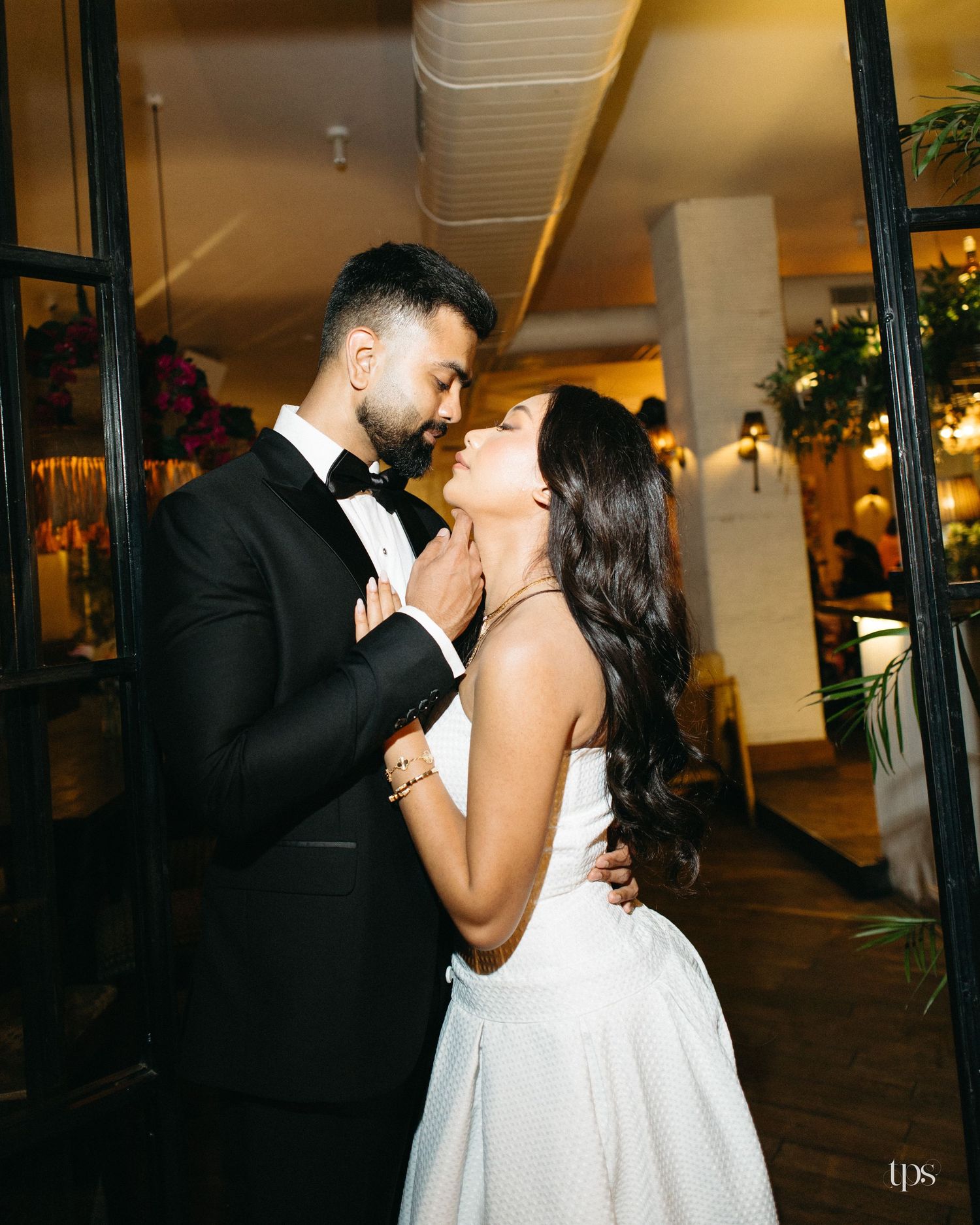 A groom in a black tuxedo and bow tie embraces his bride in a white dress as she tilts her chin up in an elegant restaurant venue.