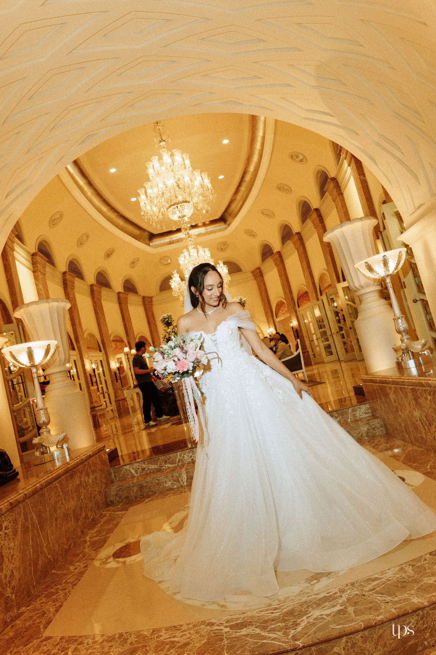 Bride in a white off-shoulder ball gown holds a bouquet in an ornate golden ballroom with chandeliers.
