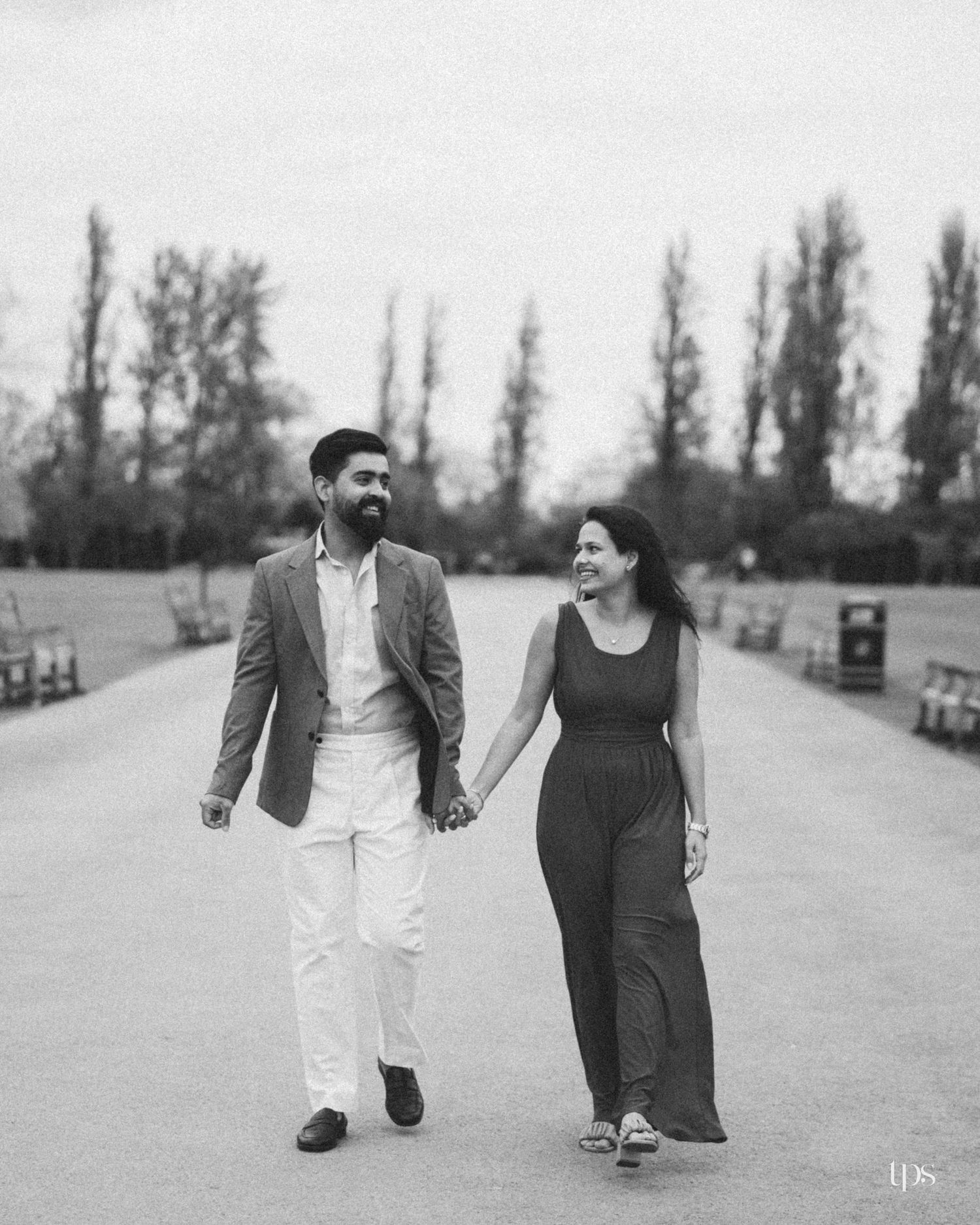 Black and white photo of a smiling couple holding hands and walking together in a park lined with tall trees.