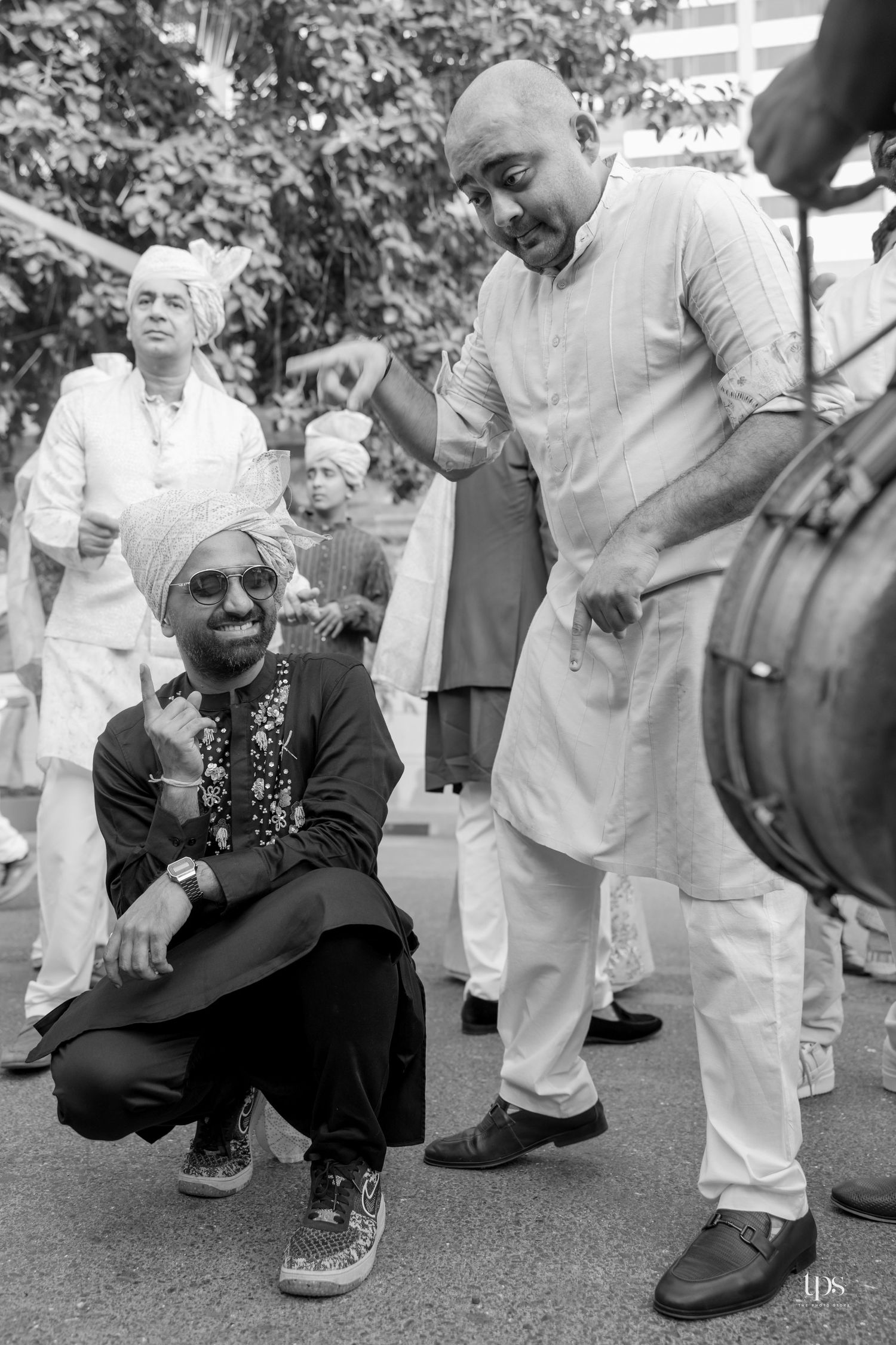 Black and white photo of men celebrating at an Indian wedding baraat, with groom in turban crouching and smiling.