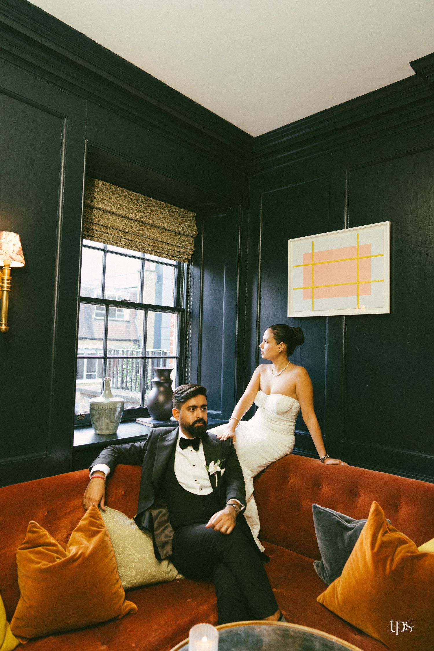 Bride and groom pose elegantly in a dark-walled room with an orange velvet sofa and modern art.