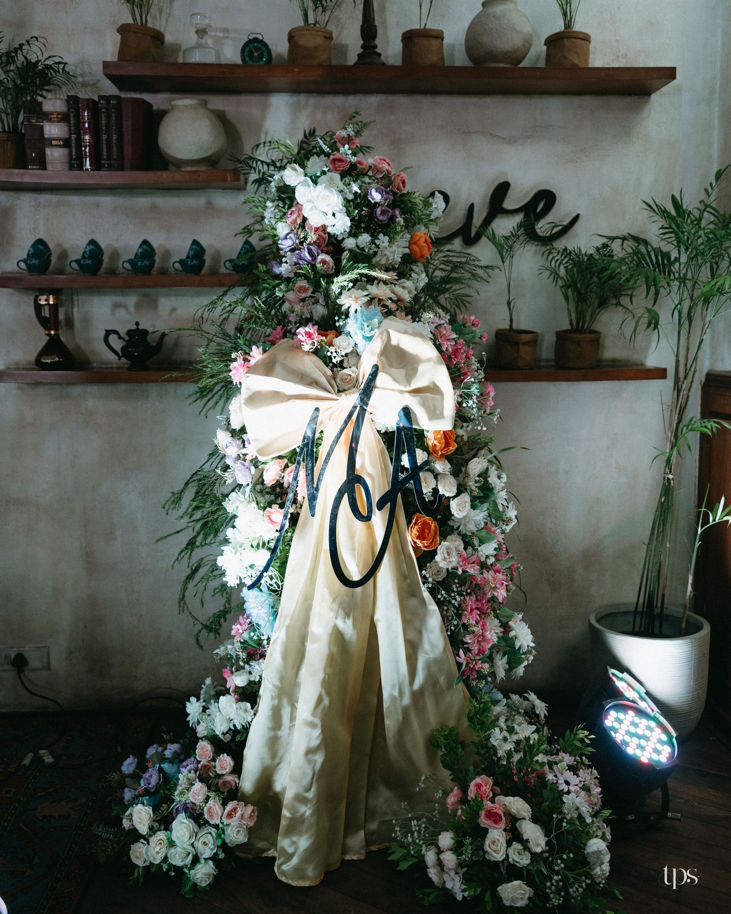 Floral standing wreath of white, pink and orange roses with a cream satin bow and ribbon, beside potted plants and shelves of teapots.
