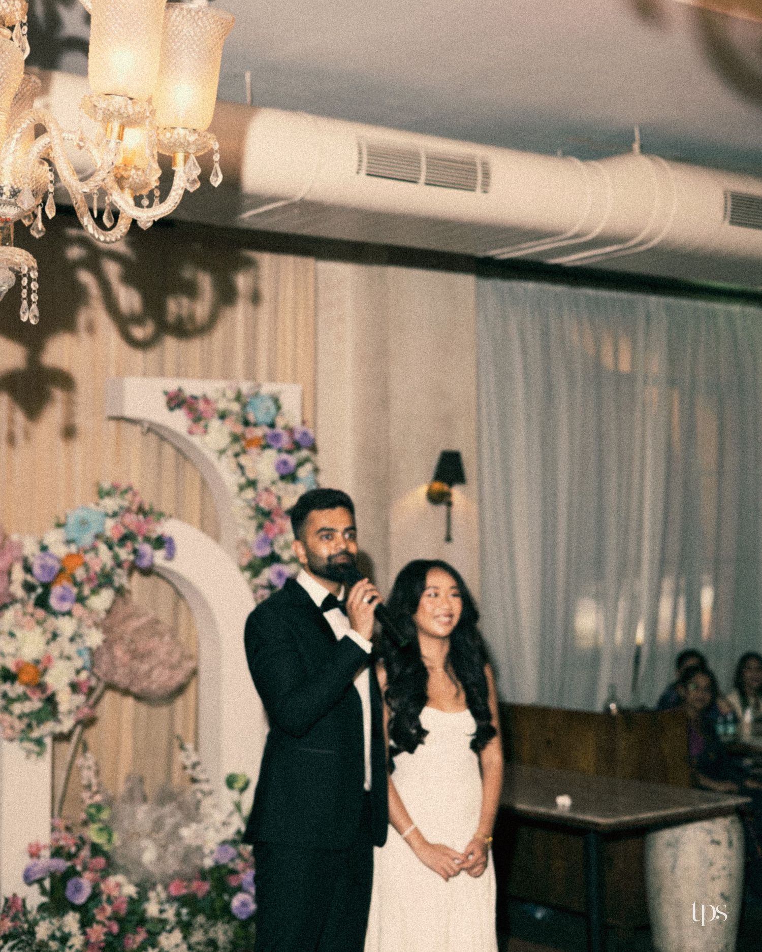 Groom in a black tuxedo speaks into a microphone beside his bride in a white dress, near a floral arch at an elegant wedding reception.