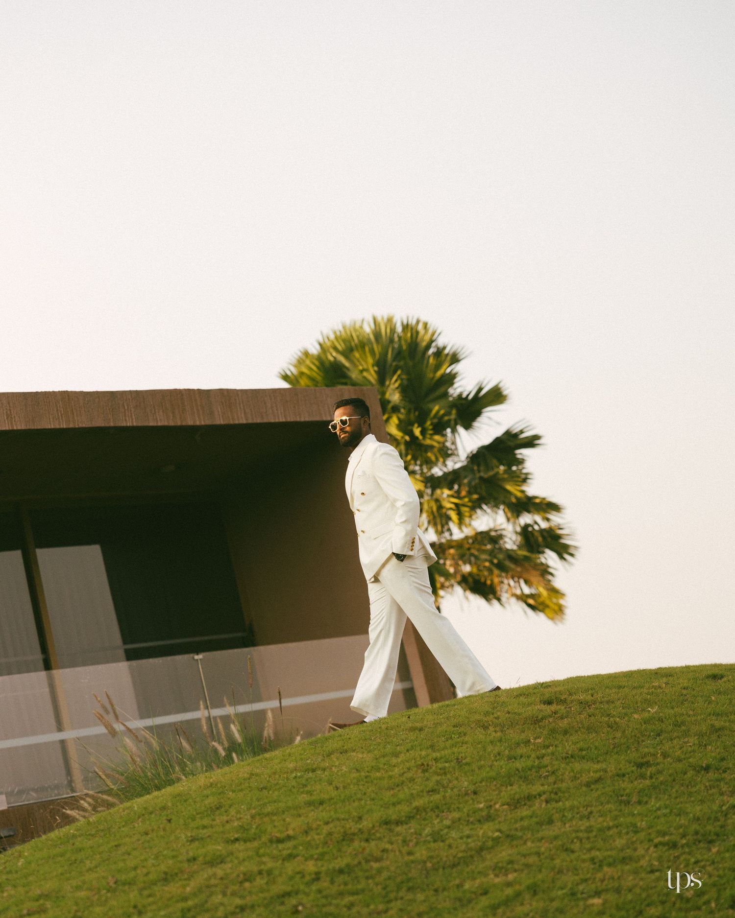 Man in a white suit and sunglasses walks along a green grassy hillside beside a modern building with a palm tree against a pale sky.