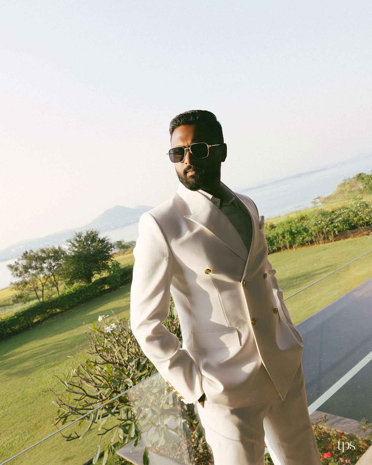 Man in a white double-breasted suit and sunglasses stands on a terrace overlooking a green lawn, trees, and hazy hills.