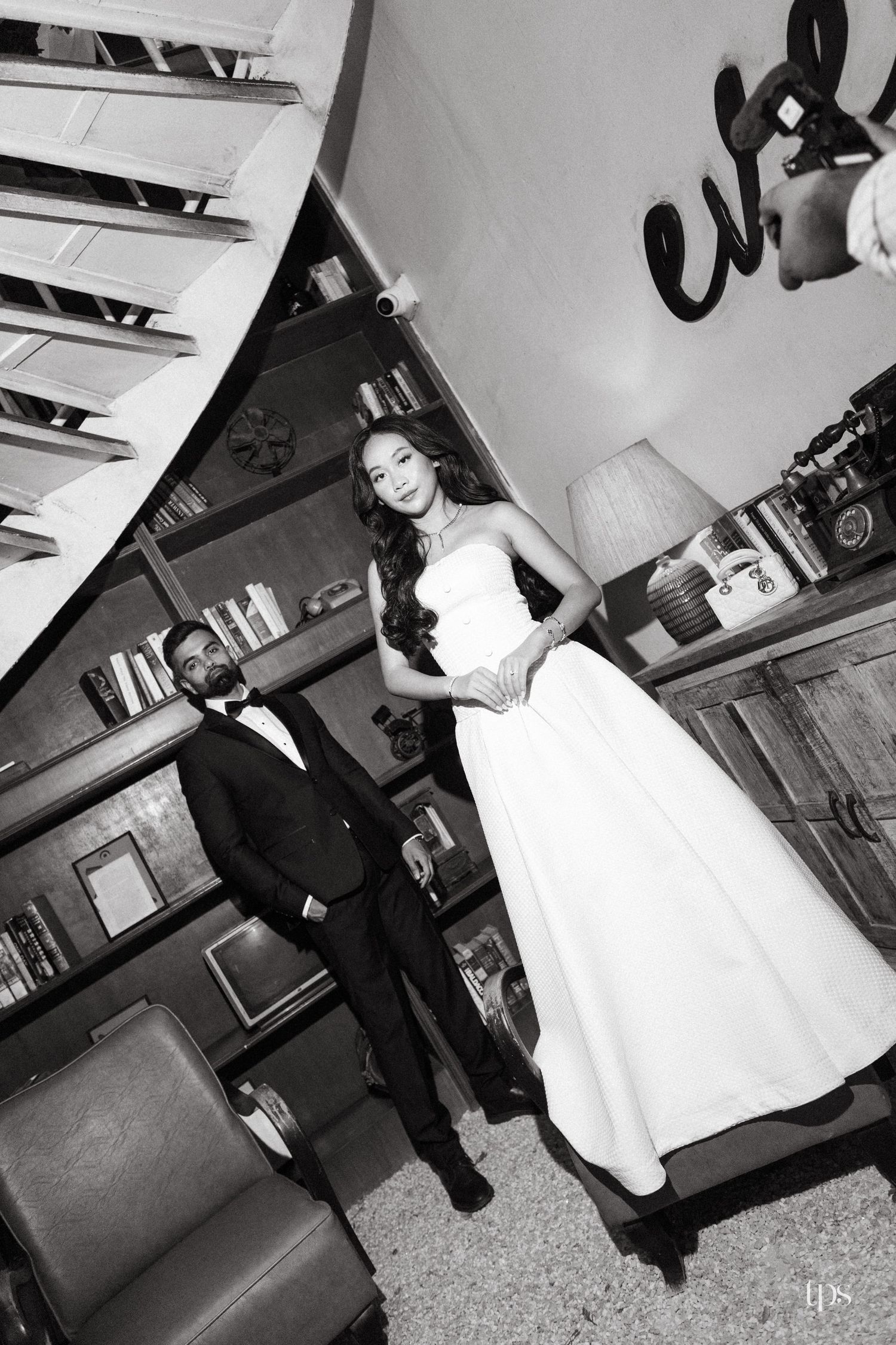 Black and white pre-wedding photo of a bride in a strapless gown and a groom in a suit posing in a cozy library with bookshelves.