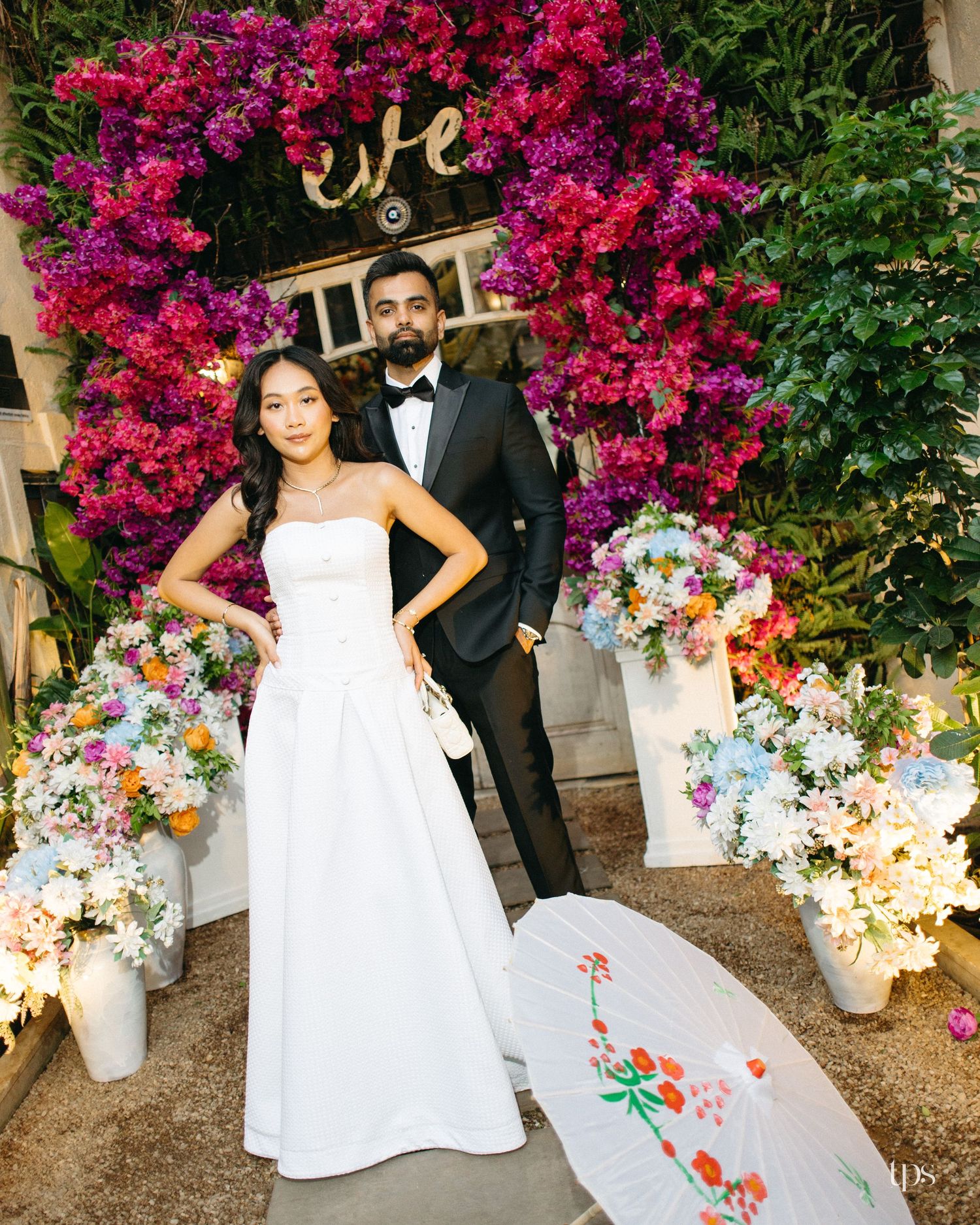 Bride in a white strapless gown and groom in a black tuxedo pose before a bougainvillea arch with floral arrangements and a paper parasol.