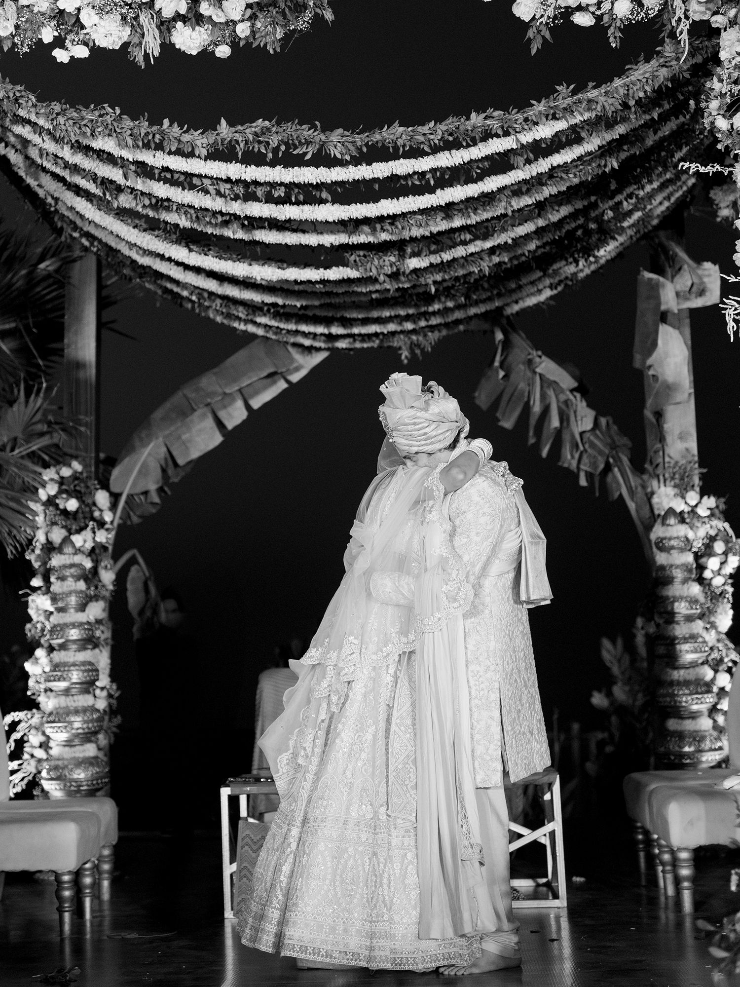 Groom in traditional Indian wedding attire stands under floral mandap decoration in black and white photo.