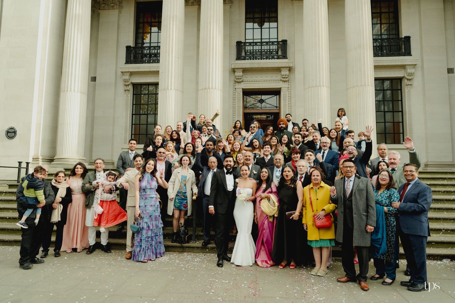 Large wedding group photo on grand building steps, bride in white gown surrounded by colorful guests celebrating.