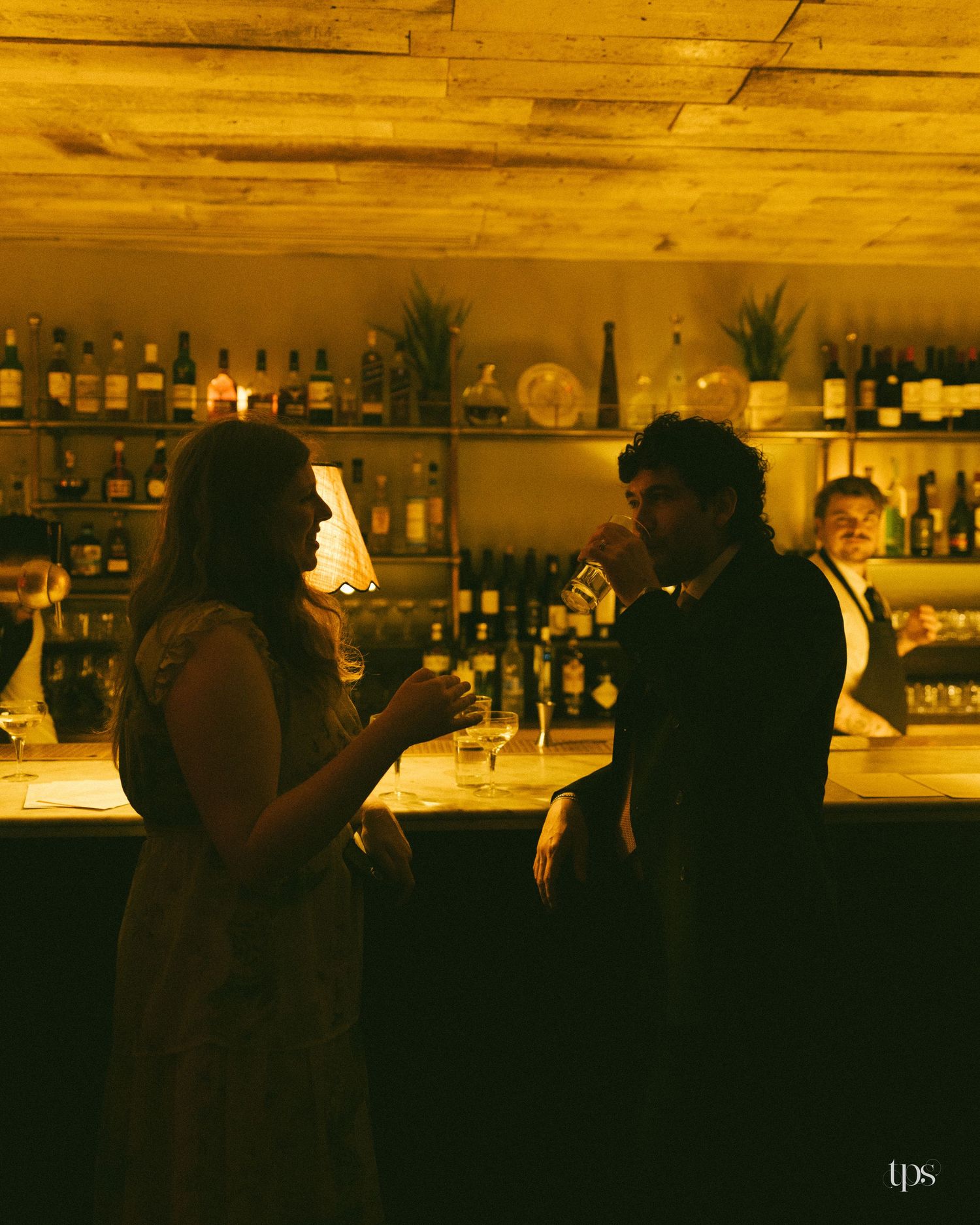 Couple sharing drinks at a warmly lit bar with wine bottles on shelves in a romantic candlelit setting.