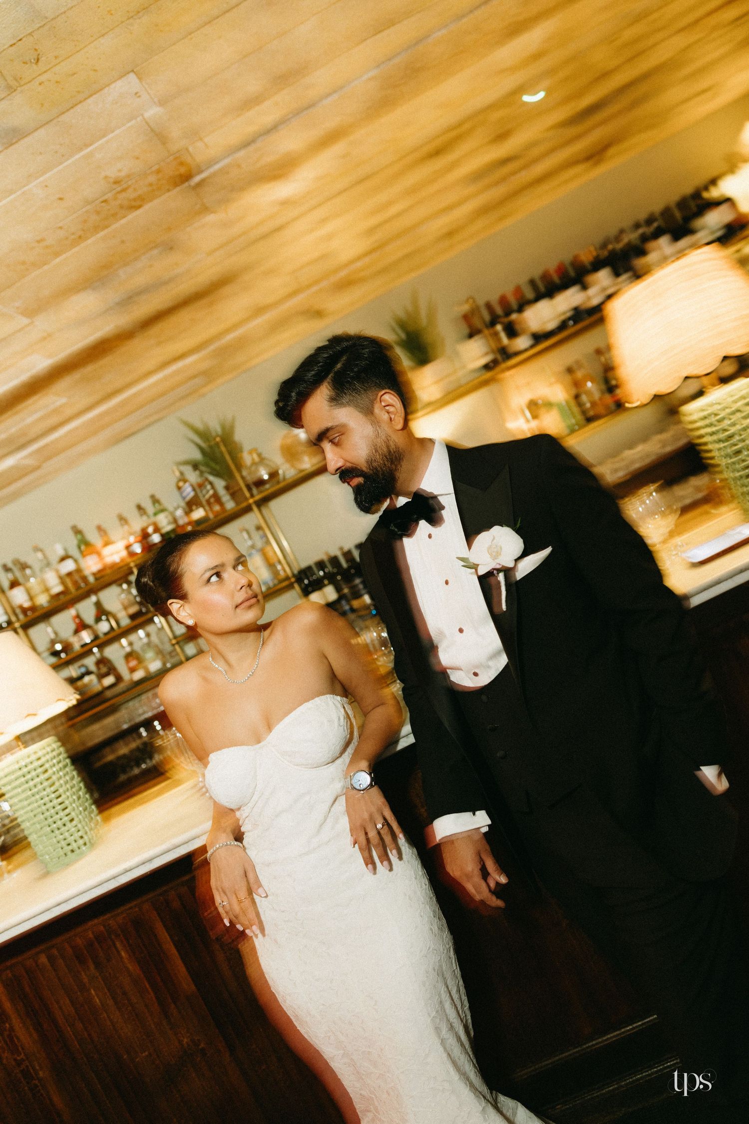 Bride in strapless white gown and groom in black tuxedo share an intimate moment at a warmly lit bar.