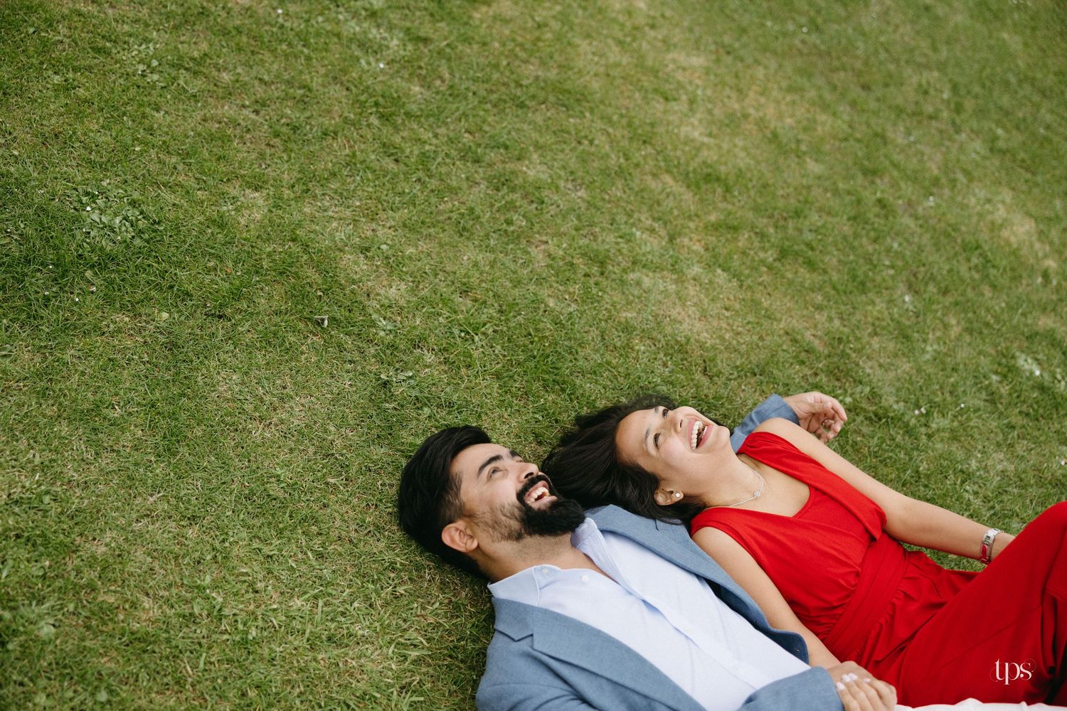 Couple laughing and lying on green grass, woman in red dress and man in blue suit, aerial view.