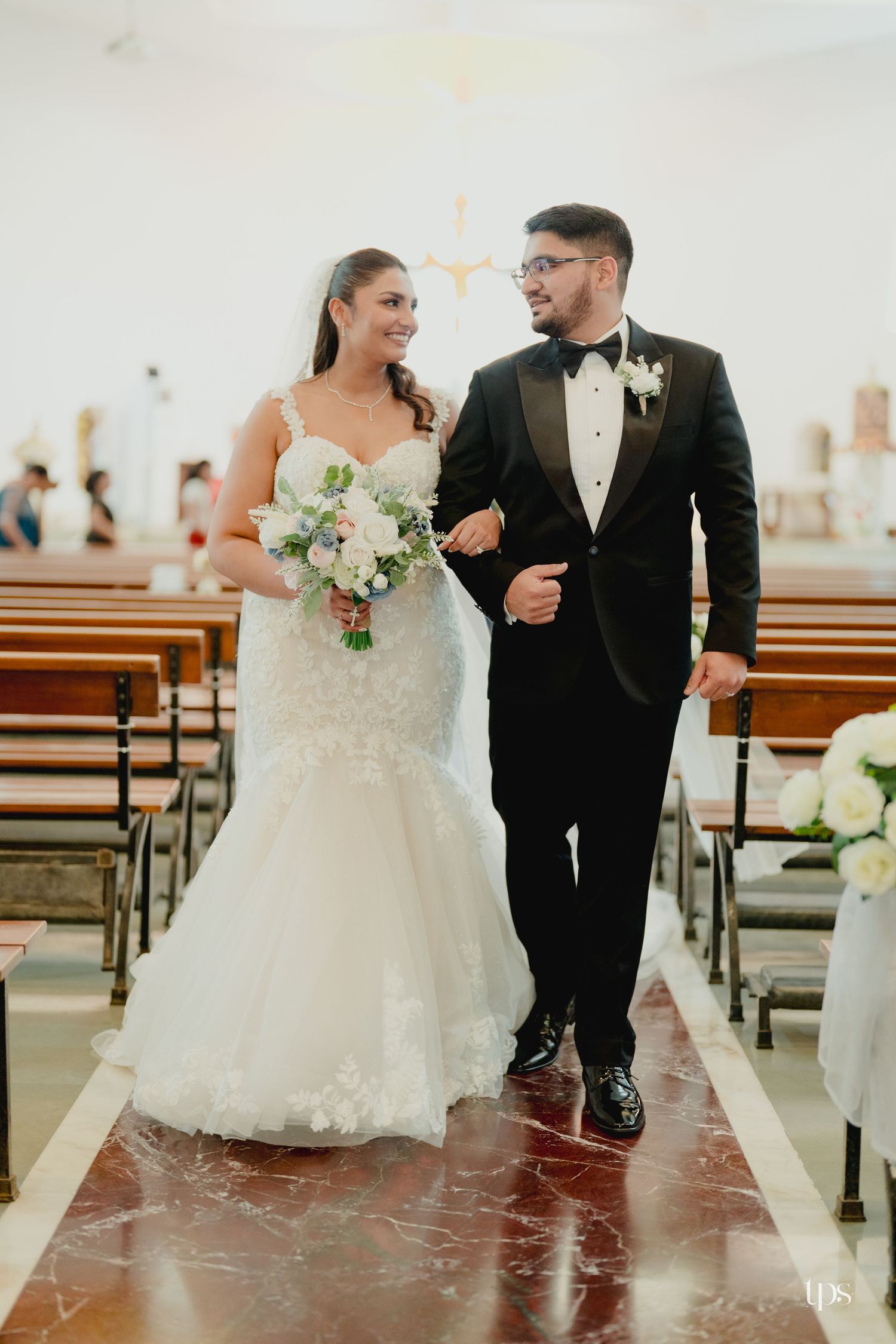 Bride in mermaid gown and groom in black tuxedo walking down church aisle after wedding ceremony.